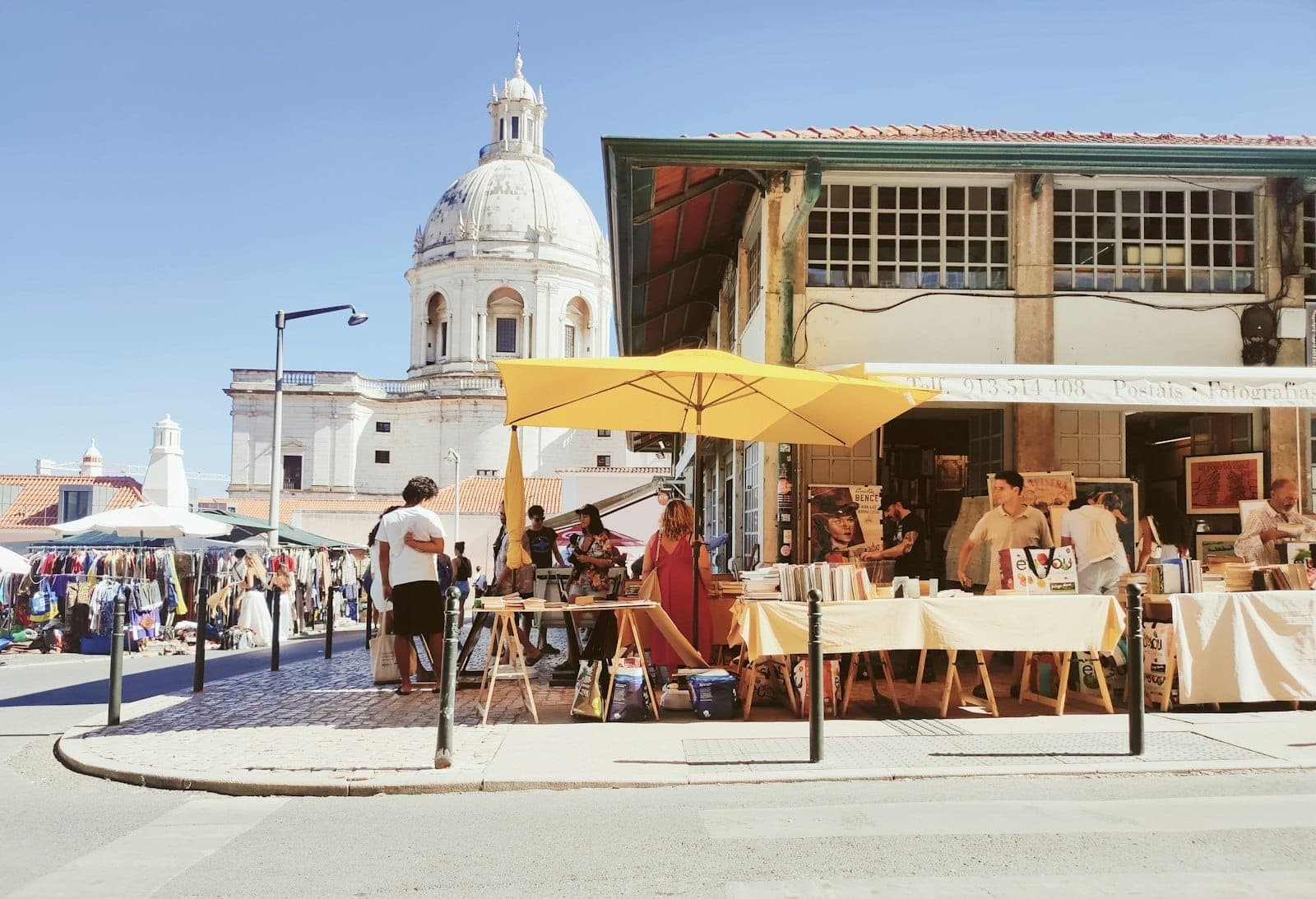 Cena de mercado de rua ao ar livre em Lisboa com barracas sob guarda-sóis amarelos, pessoas passeando e uma igreja com cúpula ao fundo.