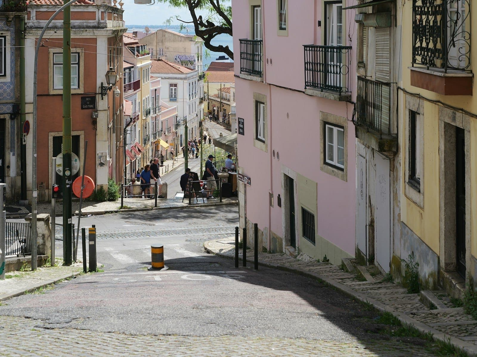 Rua de paralelepípedos tranquila em Lisboa, ladeada por casas em tons pastel, com alguns moradores caminhando e uma vista distante de colinas e telhados sob a luz suave da tarde.