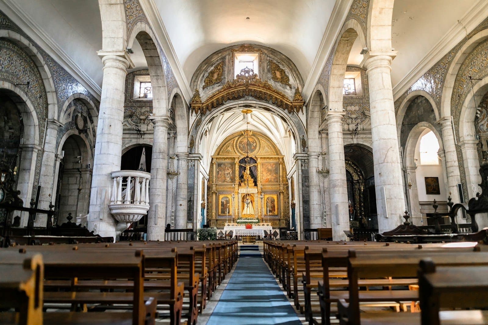 Interior bem iluminado de uma histórica igreja de pedra em Lisboa com tetos abobadados, colunas clássicas, bancos de madeira e um ornamentado retábulo dourado na frente.