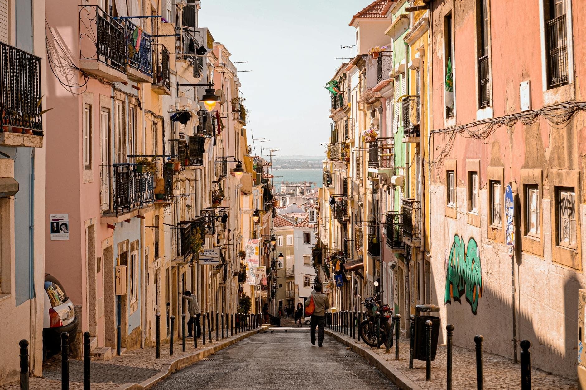 Rua íngreme e estreita ladeada por coloridos edifícios tradicionais de Lisboa, pessoas caminhando morro abaixo e vista distante da paisagem urbana e da água.