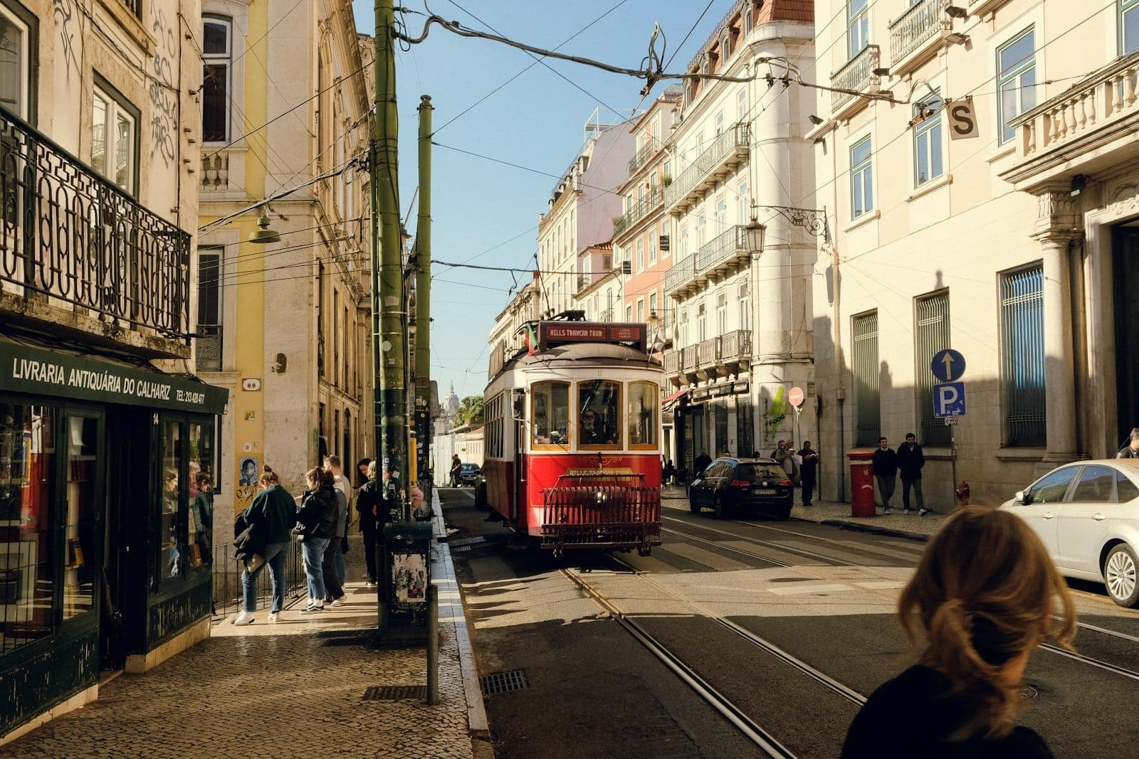 Bonde histórico de Lisboa percorrendo uma rua da cidade com pedestres, edifícios tradicionais e fios aéreos do elétrico visíveis.