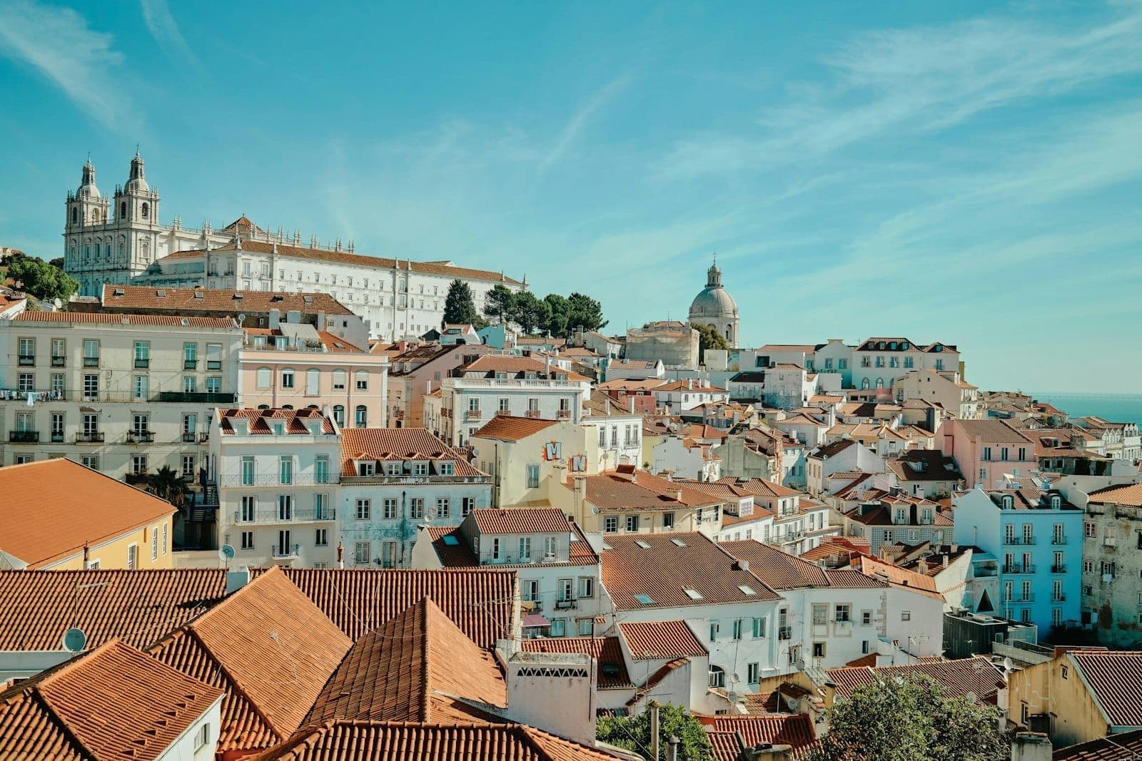 Vista panorâmica do bairro da Alfama, em Lisboa, com seus icônicos telhados vermelhos, prédios históricos e o Rio Tejo sob um céu azul brilhante com nuvens leves.