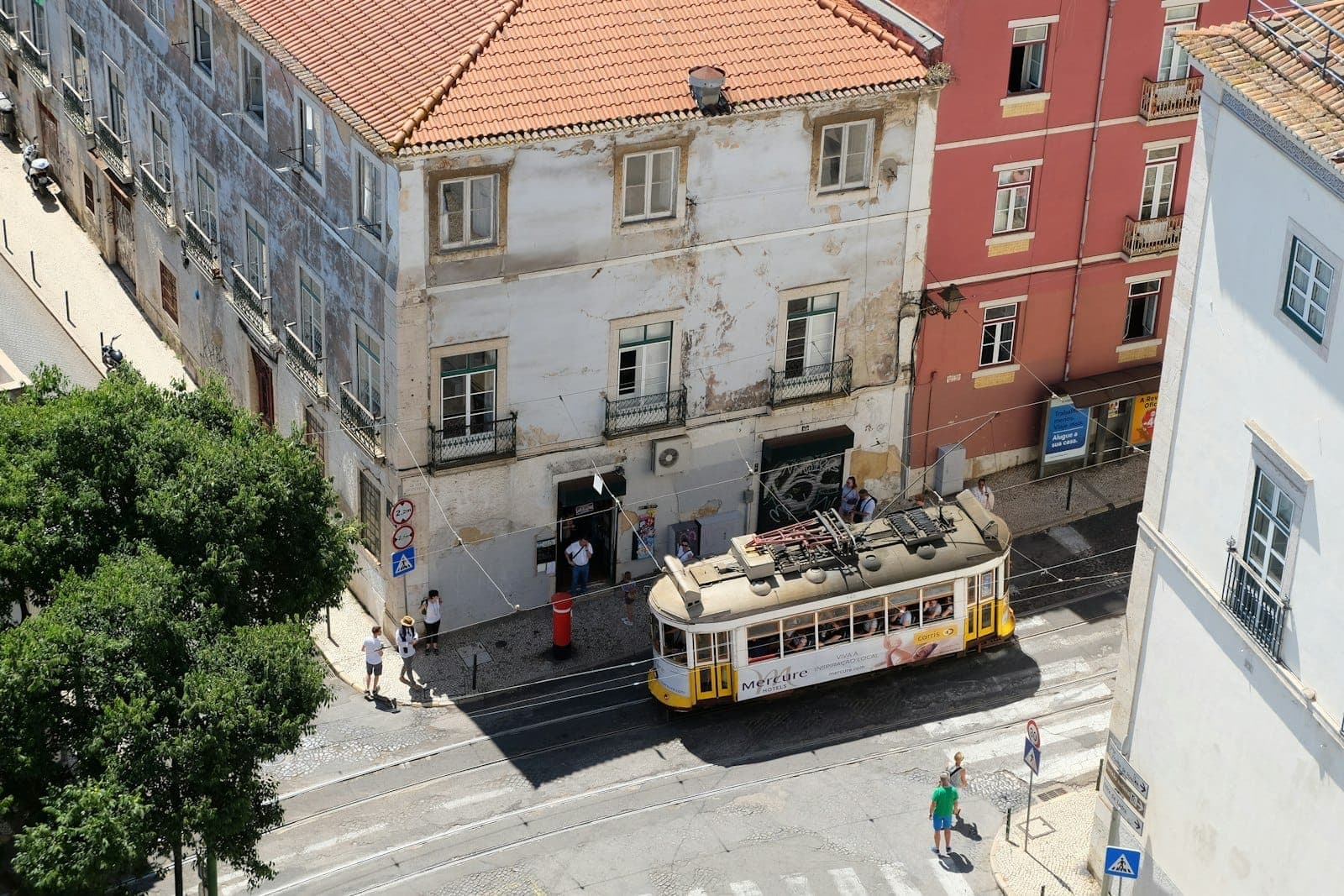 Um histórico bonde amarelo dobra uma esquina no bairro de Alfama em Lisboa, passando por antigos edifícios residenciais com telhados de telhas e pessoas caminhando.