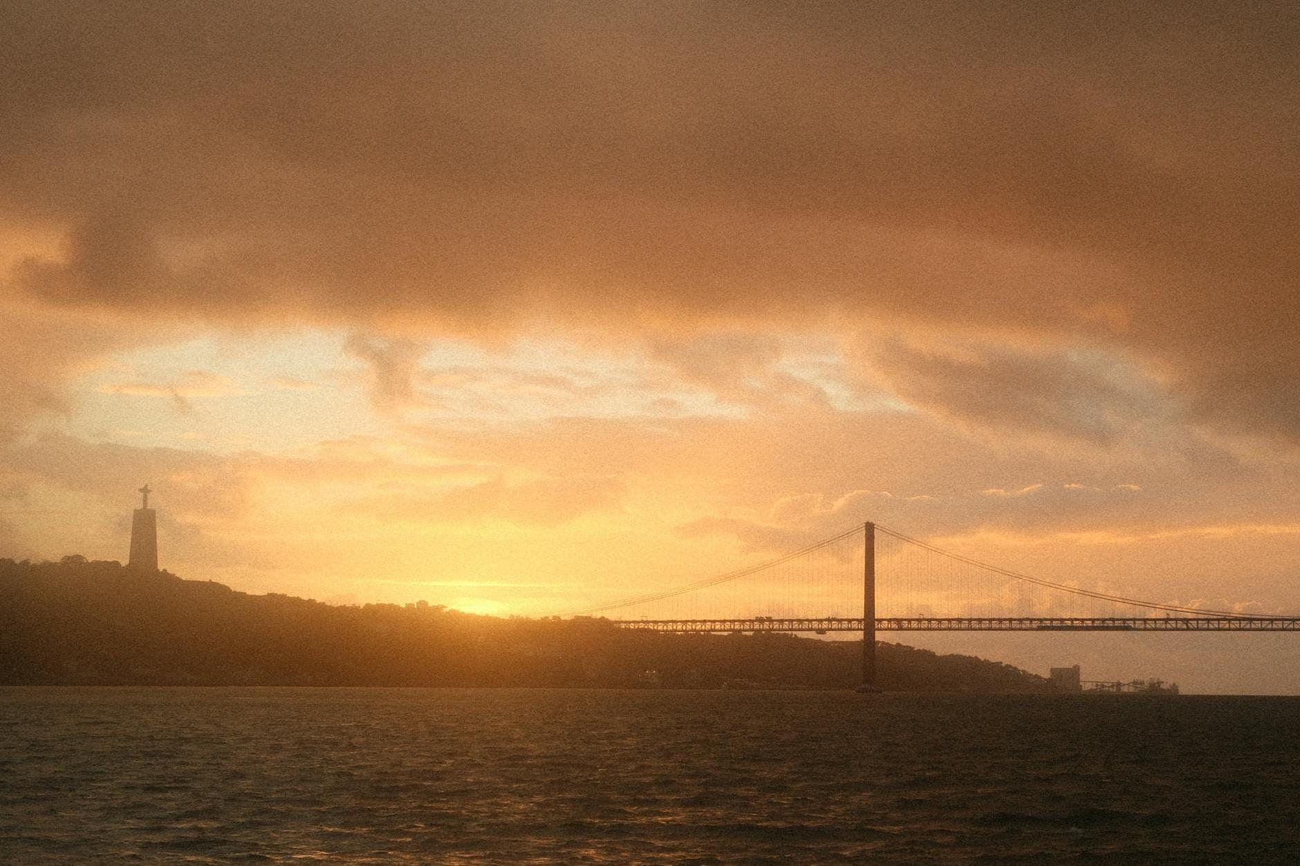 A Ponte 25 de Abril de Lisboa ao pôr do sol dourado, com o rio Tejo em primeiro plano e a estátua do Cristo Rei visível nas colinas.