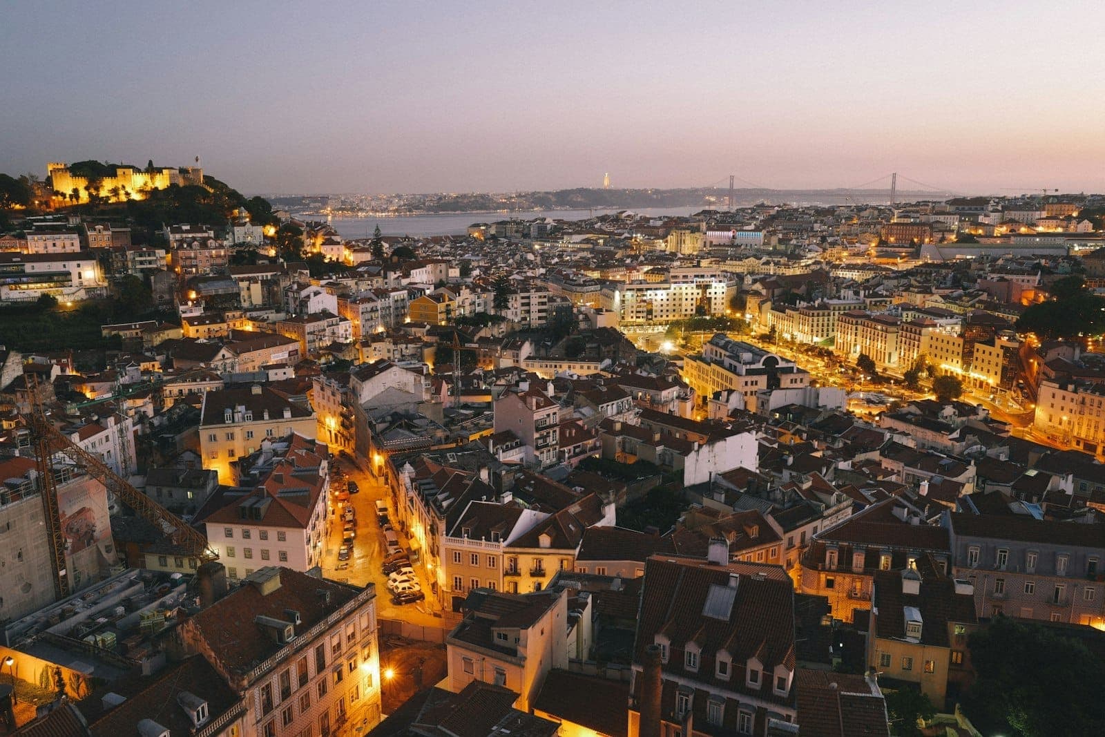 Panorama dramático de Lisboa ao entardecer com luzes quentes da cidade, o castelo iluminado, ruas sinuosas e o rio ao fundo, transmitindo toda a energia da noite lisboeta.