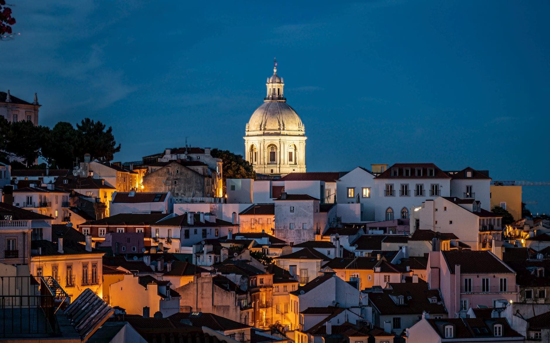 O bairro de Alfama em Lisboa à noite com cúpula iluminada, edifícios caiados de branco e telhados cor de laranja sob um céu azul profundo.