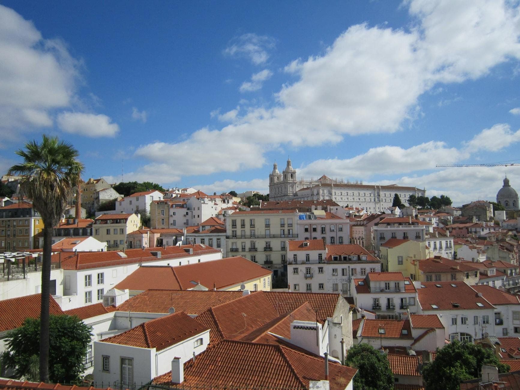 Vista panorâmica sobre os telhados do bairro de Alfama em Lisboa com edifícios históricos, igrejas e céu azul, mostrando as belas paisagens urbanas típicas dos miradouros gratuitos ou de baixo custo.