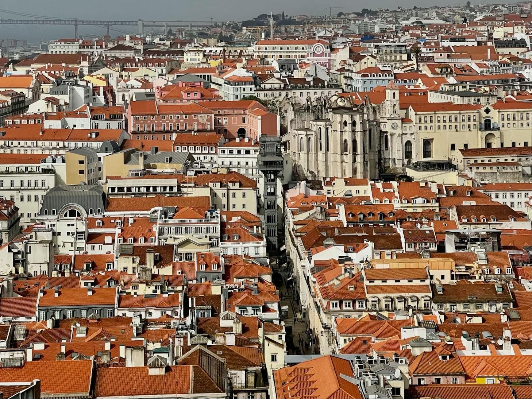 Vista aérea do centro de Lisboa, mostrando telhados de telhas vermelhas, vários bairros, edifícios históricos e o rio ao fundo