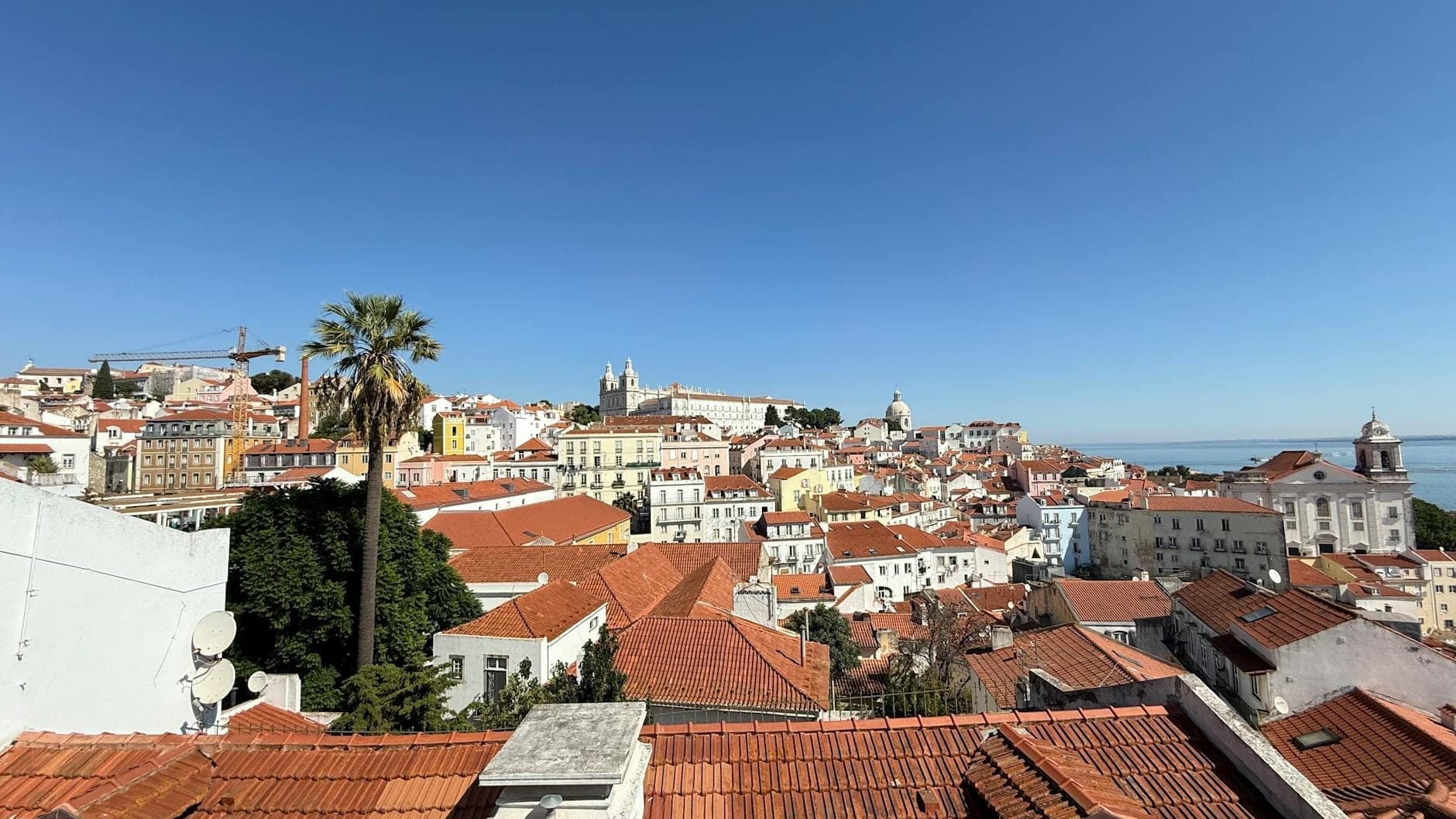 Vista panorâmica dos bairros históricos de Lisboa com telhados vermelhos, edifícios brancos, pontos turísticos marcantes e o rio ao fundo em um dia ensolarado.