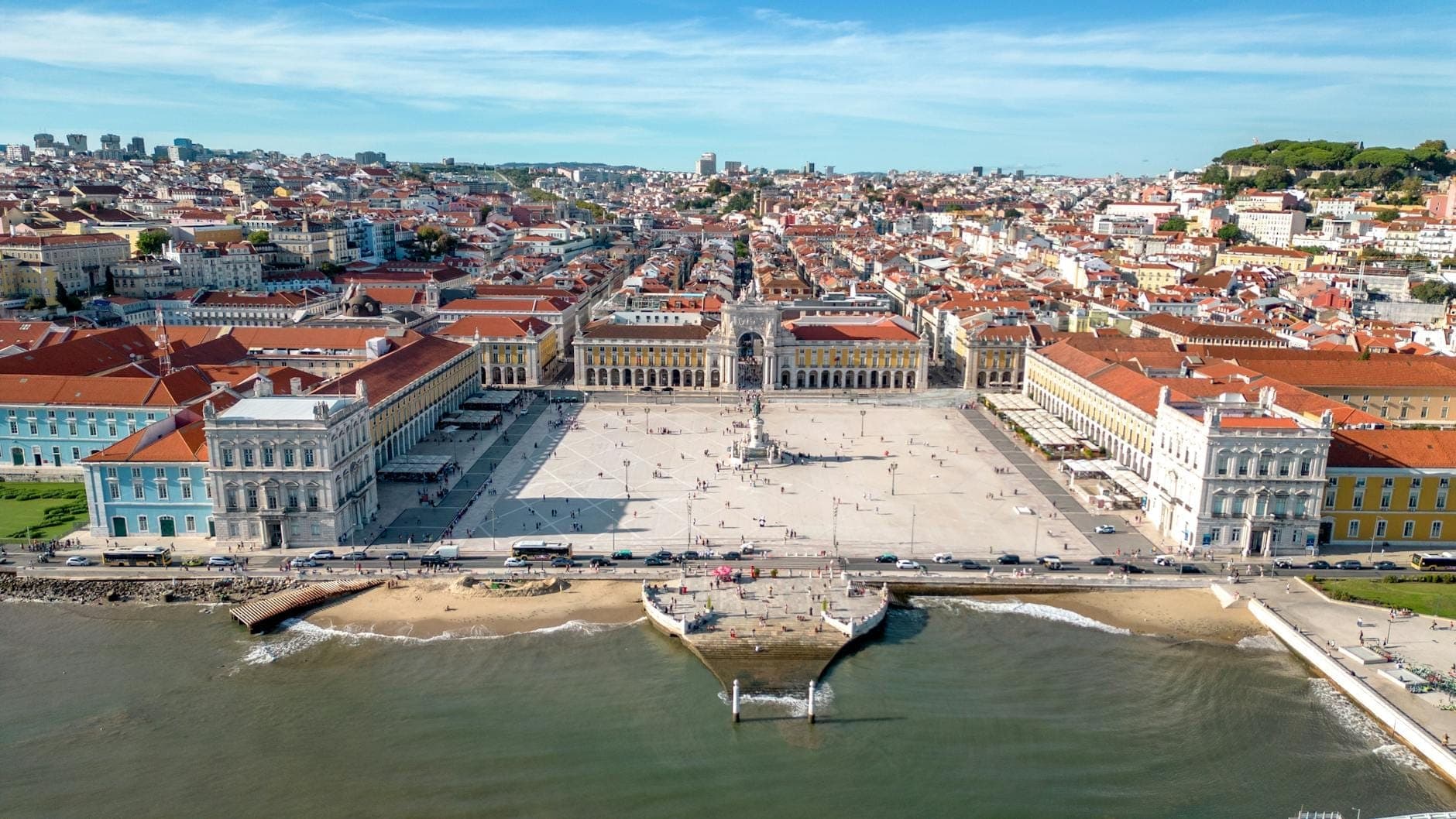 Vista aérea da Praça do Comércio em Lisboa com a margem do rio e os edifícios da cidade visíveis atrás da praça.