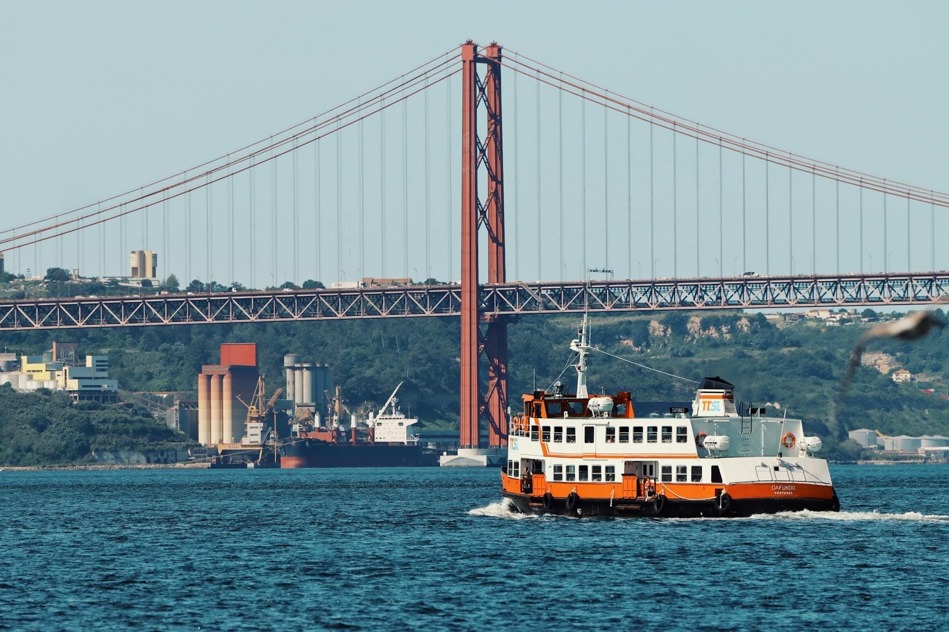 Barco clássico navegando pelo Rio Tejo em Lisboa com a Ponte 25 de Abril ao fundo em um dia ensolarado.