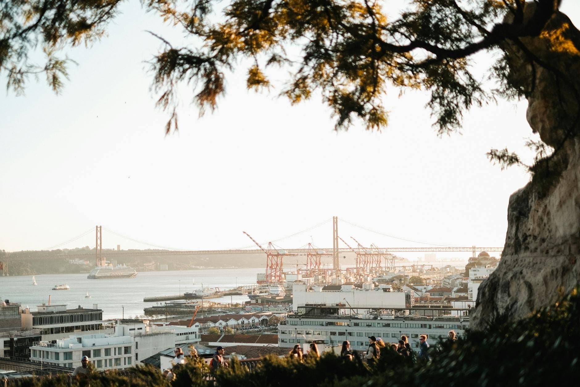 Multidões reunidas num miradouro de Lisboa com vista para o rio Tejo, com a Ponte 25 de Abril e a luz do verão ao fundo.