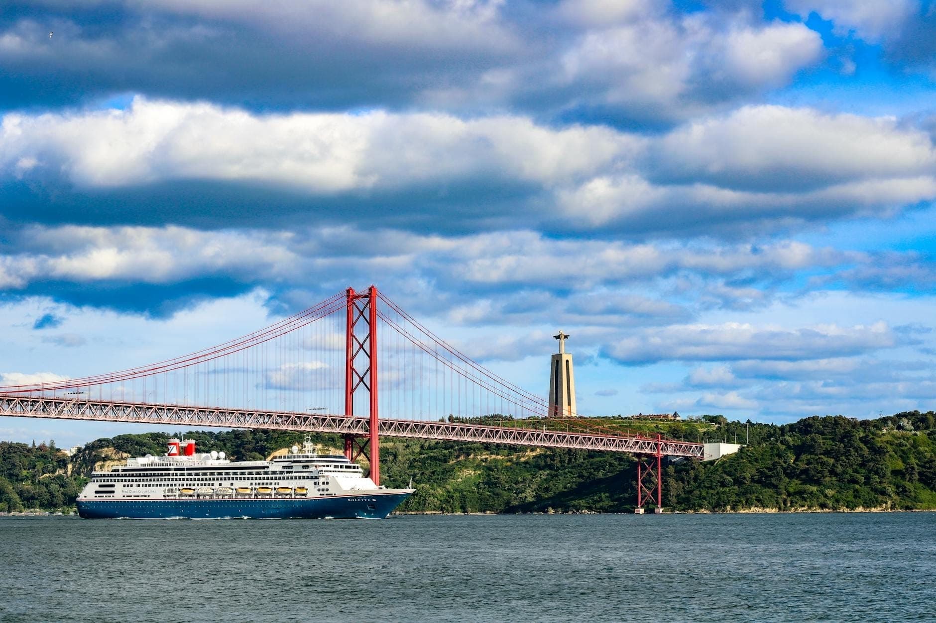 Um grande navio de cruzeiro navega sob a Ponte 25 de Abril de Lisboa com o monumento Cristo Rei visível na margem do rio.