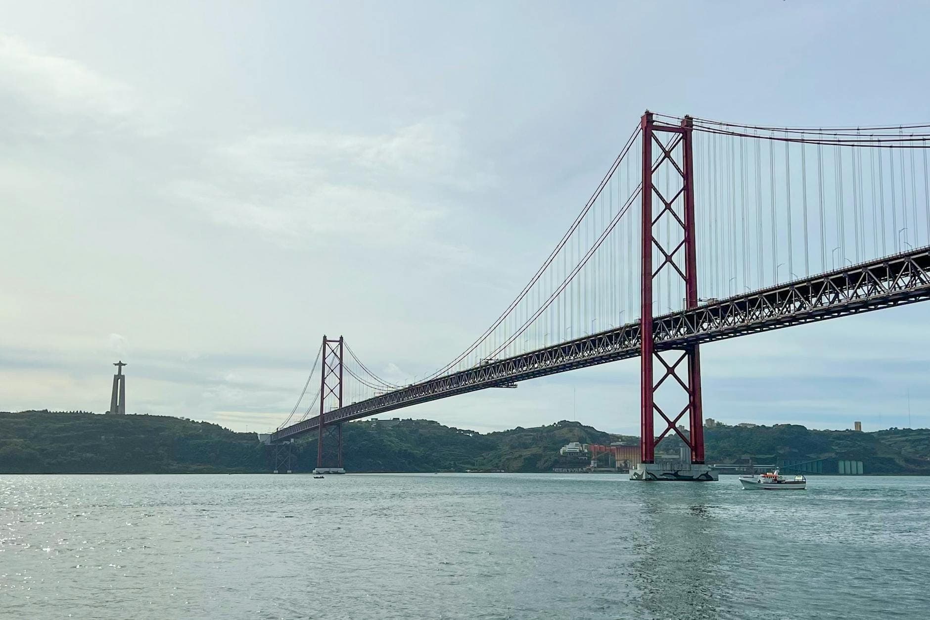 A Ponte 25 de Abril de Lisboa estendendo-se sobre o rio Tejo com a estátua do Cristo Rei visível ao fundo, vista a partir da água.