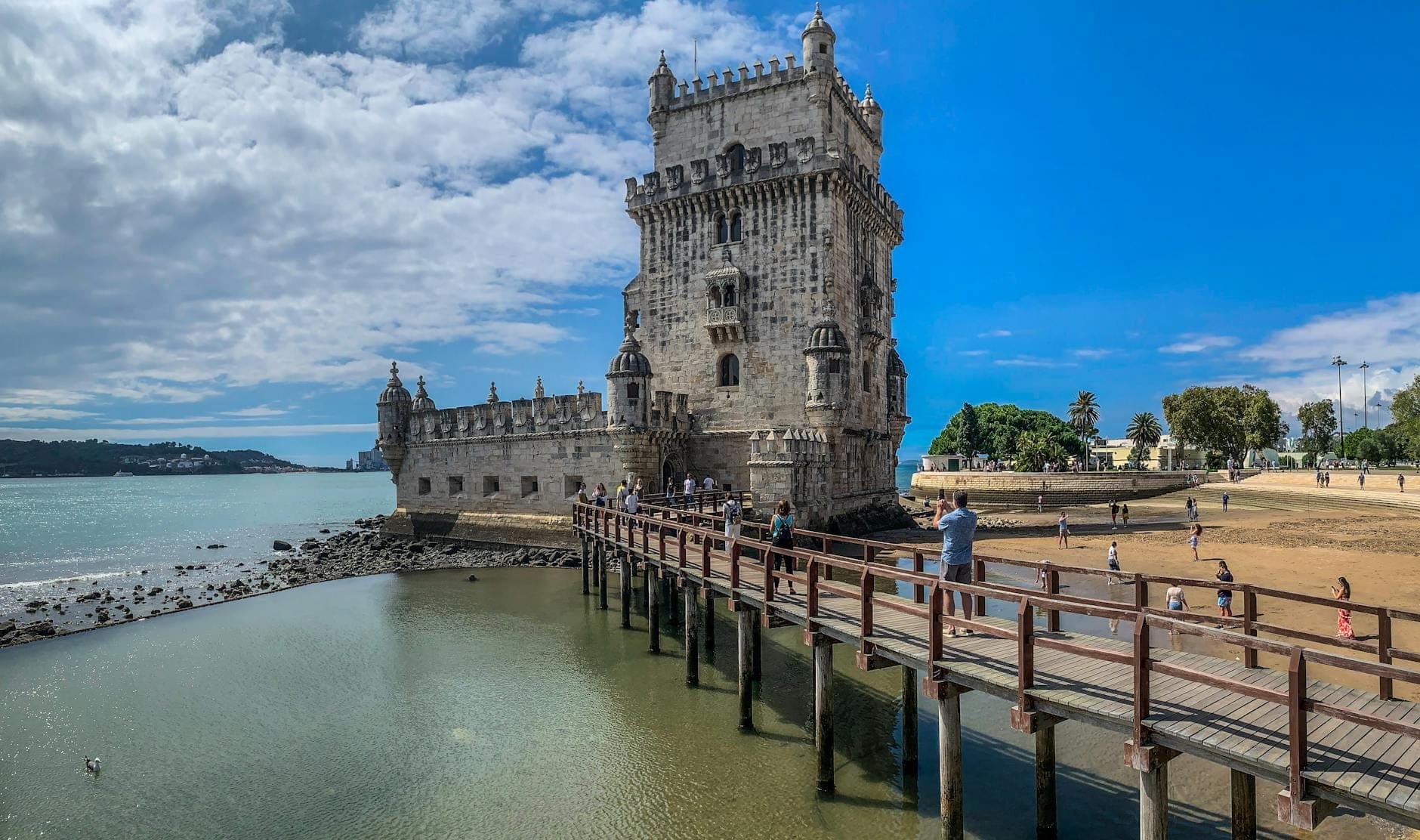 Vista ampla da Torre de Belém em Lisboa com sua passarela de madeira, orla ribeirinha e visitantes, sob um céu azul intenso.