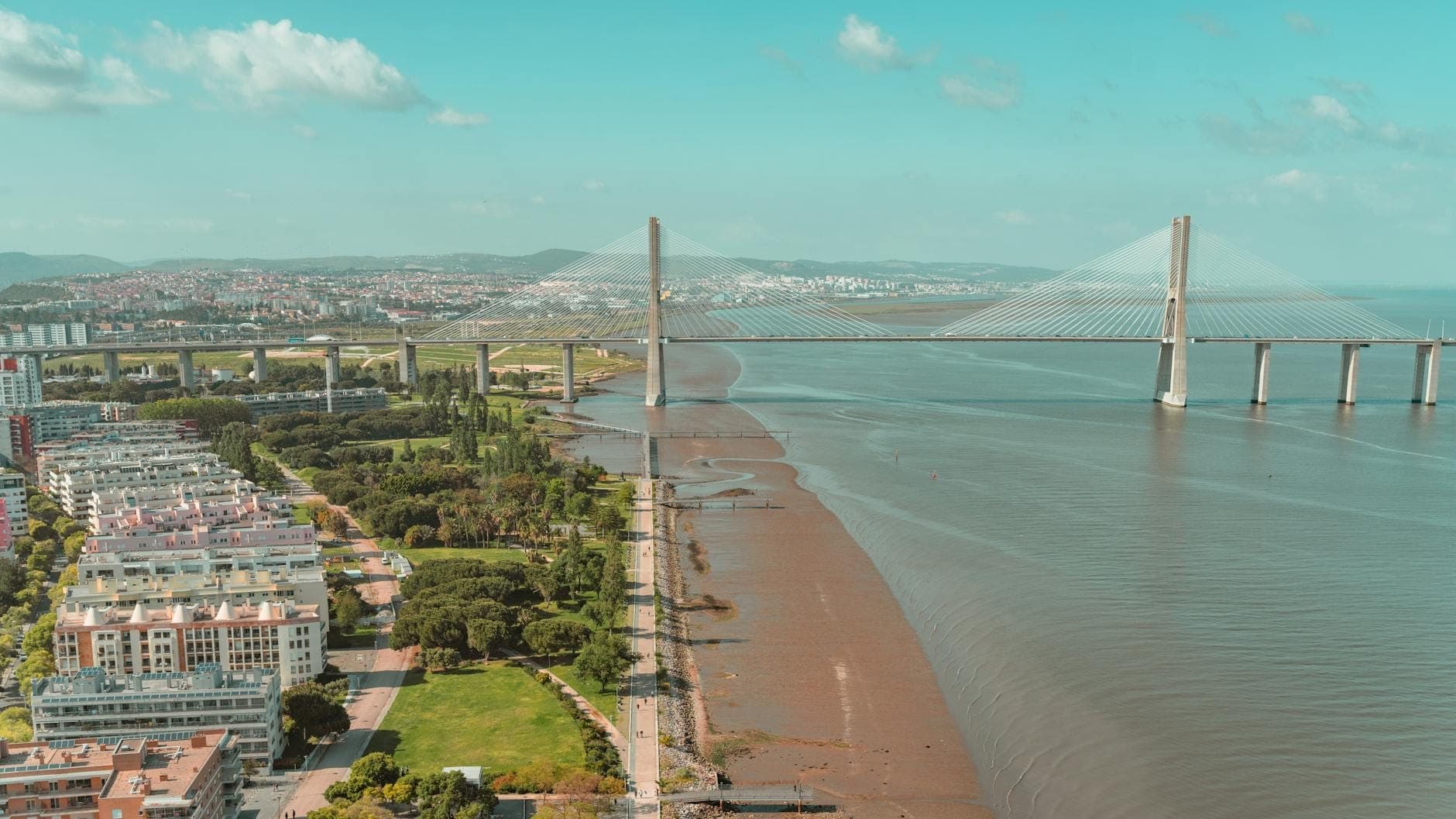 Vista aérea de Lisboa mostrando a Ponte Vasco da Gama, edifícios modernos à beira-rio e áreas verdes no bairro do Parque das Nações.