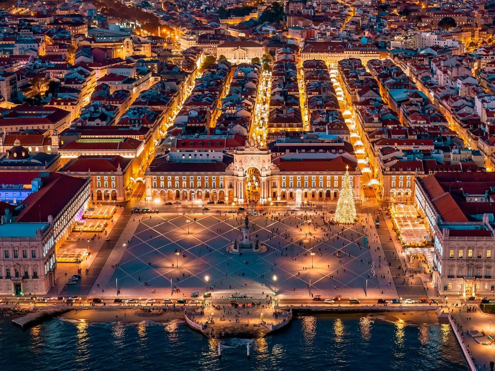Vista aérea da Praça do Comércio de Lisboa ao entardecer, com luzes da cidade, orla marítima e pessoas na praça, ilustrando um ambiente urbano seguro.