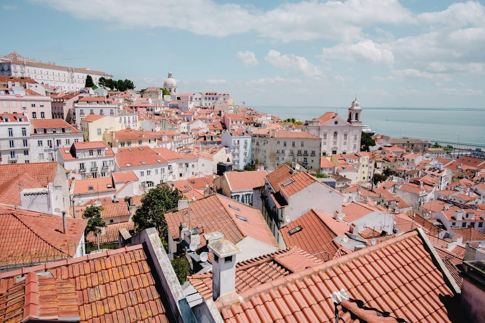 Scenic panoramic view over Lisbon’s historic Alfama district with red rooftops, pastel buildings, iconic churches, and the Tagus river under a bright, partly cloudy sky.