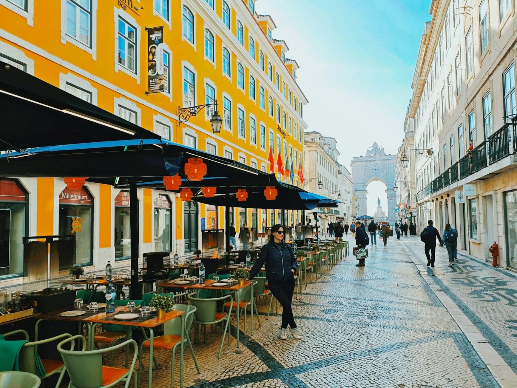 Movimentada rua comercial pedonal em Lisboa com pessoas, cafés ao ar livre, edifícios amarelos e o Arco da Rua Augusta ao fundo.