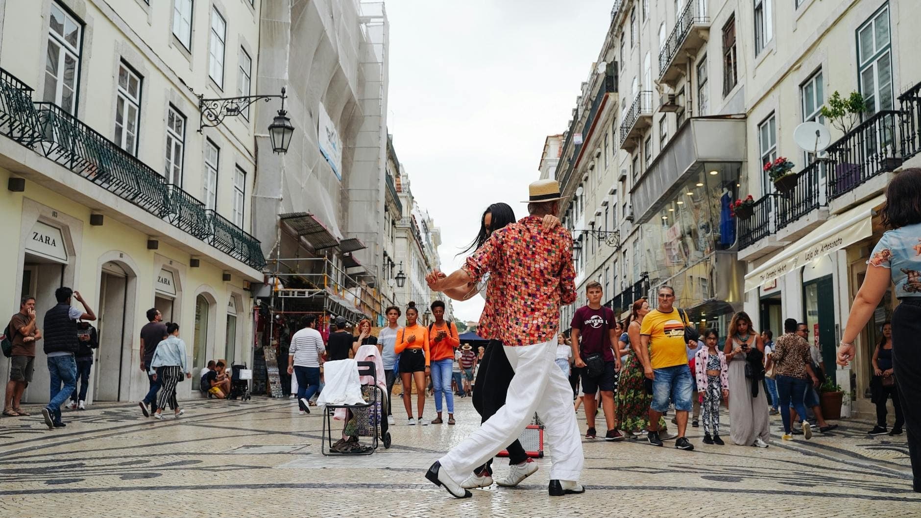 Ampla rua comercial para pedestres em Lisboa ladeada por lojas, pessoas caminhando e fazendo compras, e um casal dançando em primeiro plano.