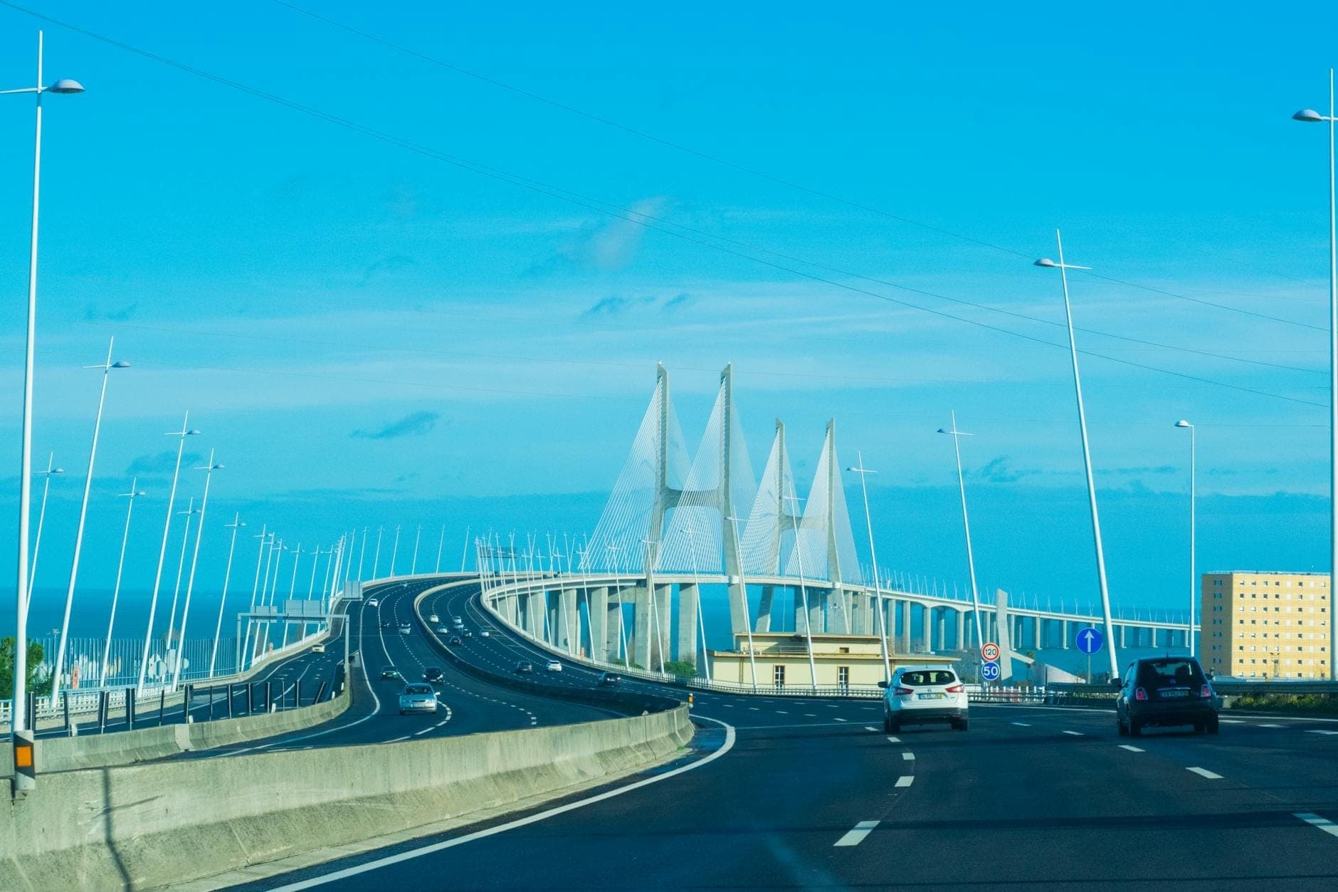 Dirigindo em uma autoestrada em Lisboa em direção à Ponte Vasco da Gama com carros, céu azul e arquitetura moderna da ponte.