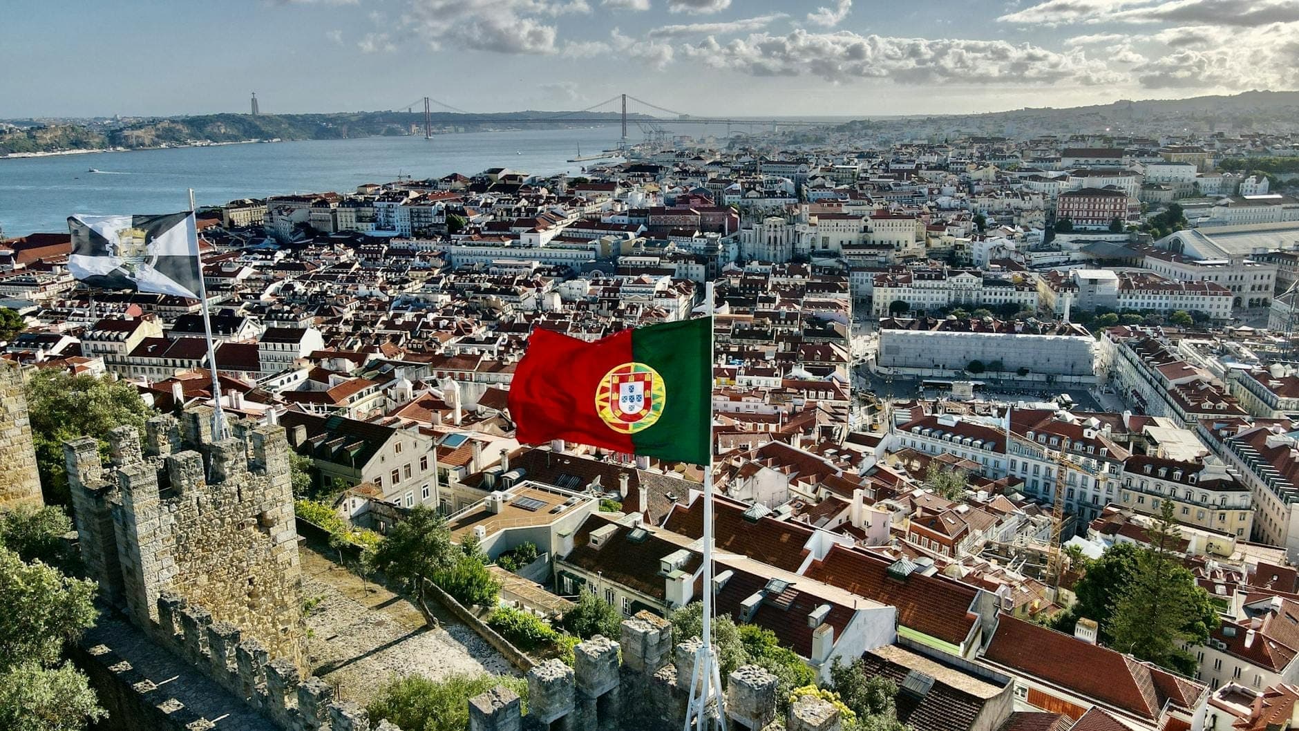 Vista aérea ampla de Lisboa com a bandeira portuguesa e os icónicos telhados vermelhos, o rio e os pontos de referência da cidade visíveis ao fundo.