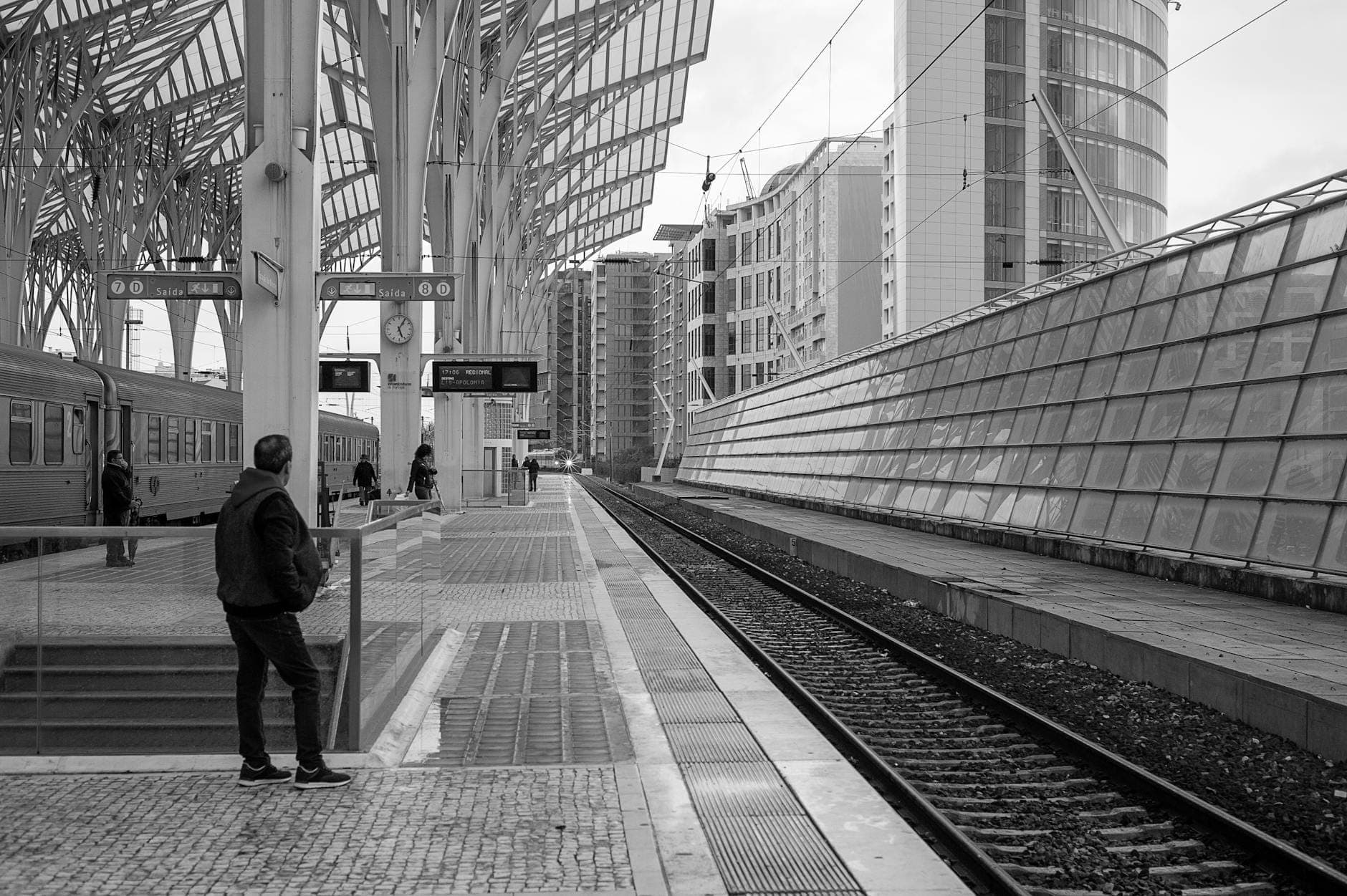 Pessoa aguardando na moderna plataforma da estação de comboios Oriente de Lisboa com trilhos e uma marcante cobertura de aço acima.