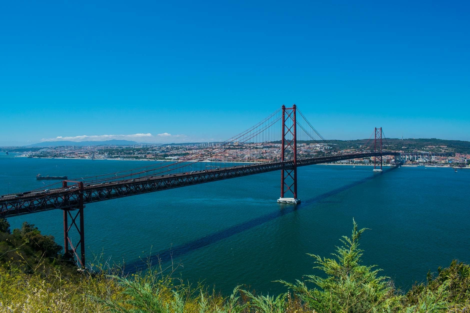 Vista ampla da Ponte 25 de Abril de Lisboa atravessando o rio Tejo, com paisagem urbana e litoral distante sob um céu azul limpo.