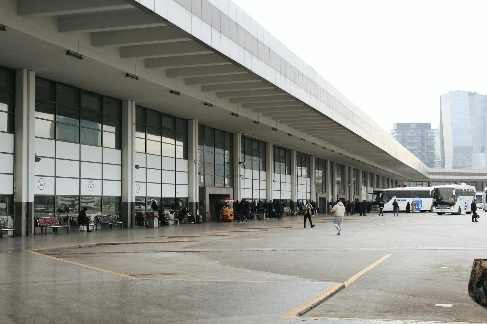 Grande terminal de ônibus moderno com pessoas esperando, ônibus estacionados e uma paisagem urbana com edifícios altos ao fundo.
