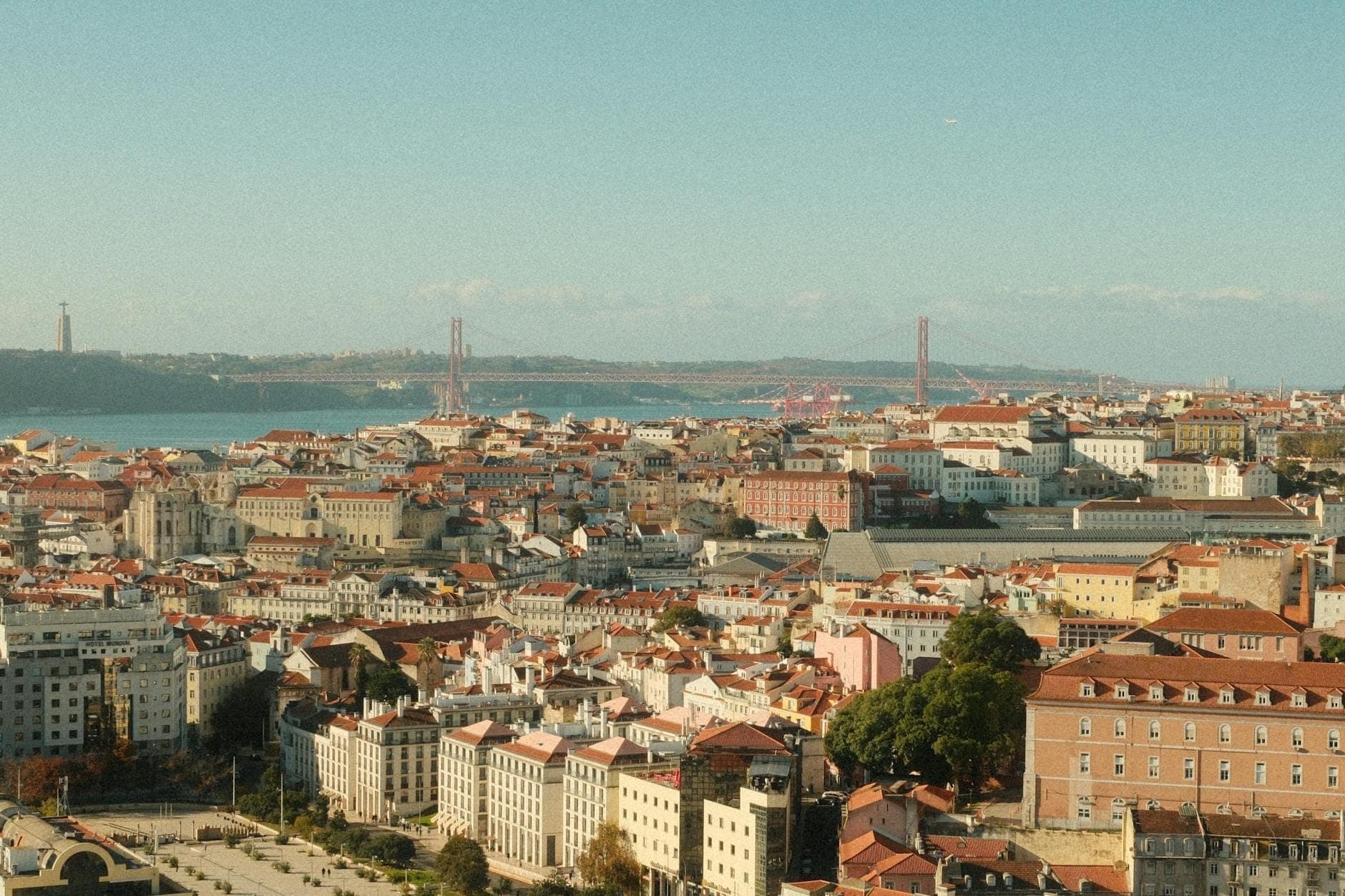 Vista panorâmica de Lisboa com telhados vermelhos, o rio Tejo e a Ponte 25 de Abril visíveis ao fundo sob um céu azul limpo.