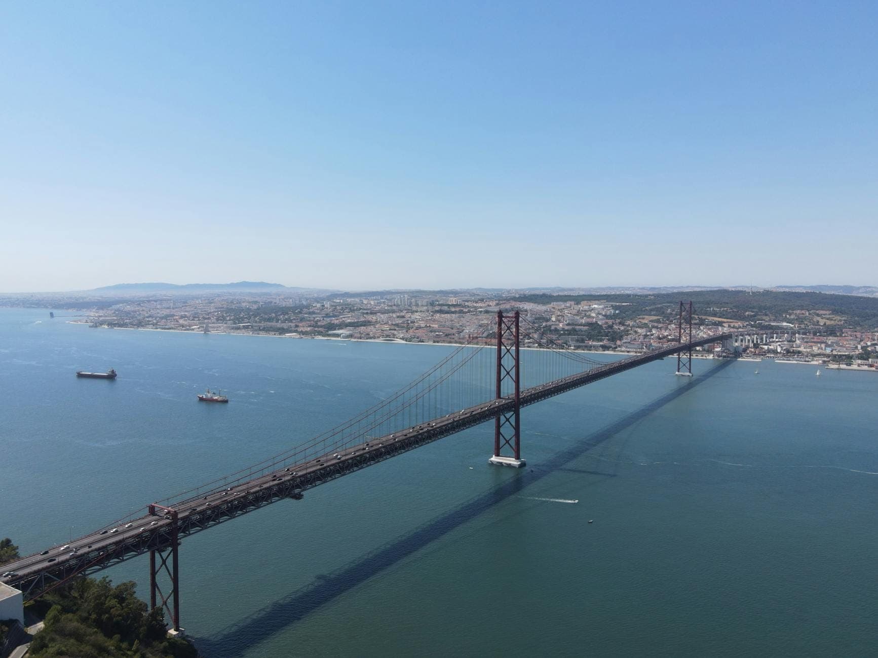 Vista aérea ampla da Ponte 25 de Abril em Lisboa sobre o rio Tejo com a cidade ao fundo e céu limpo.