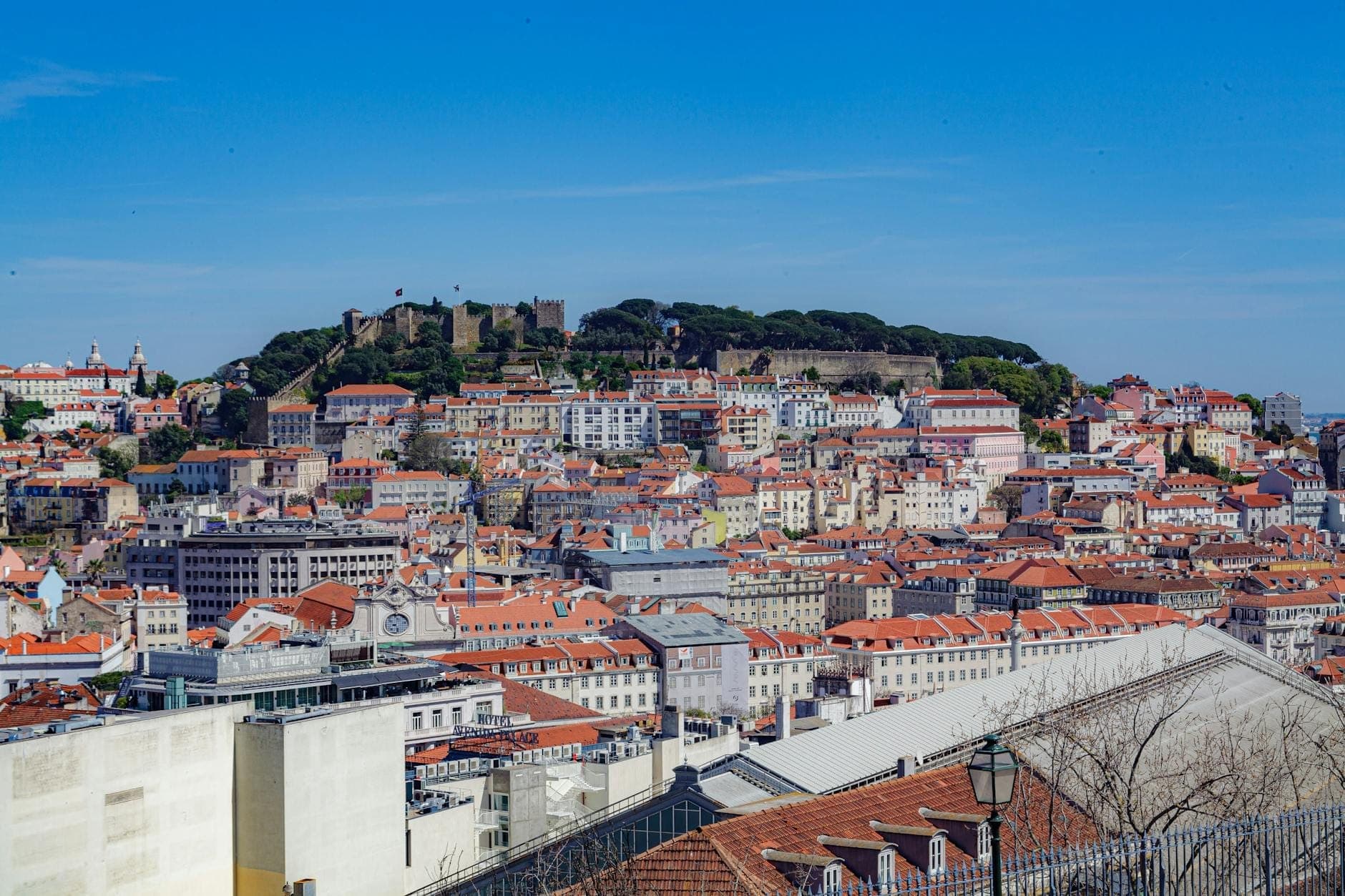 Vista panorâmica de Lisboa com um castelo histórico no alto de uma colina, edifícios coloridos com telhados vermelhos, mostrando o traçado acidentado da cidade sob um céu azul limpo.