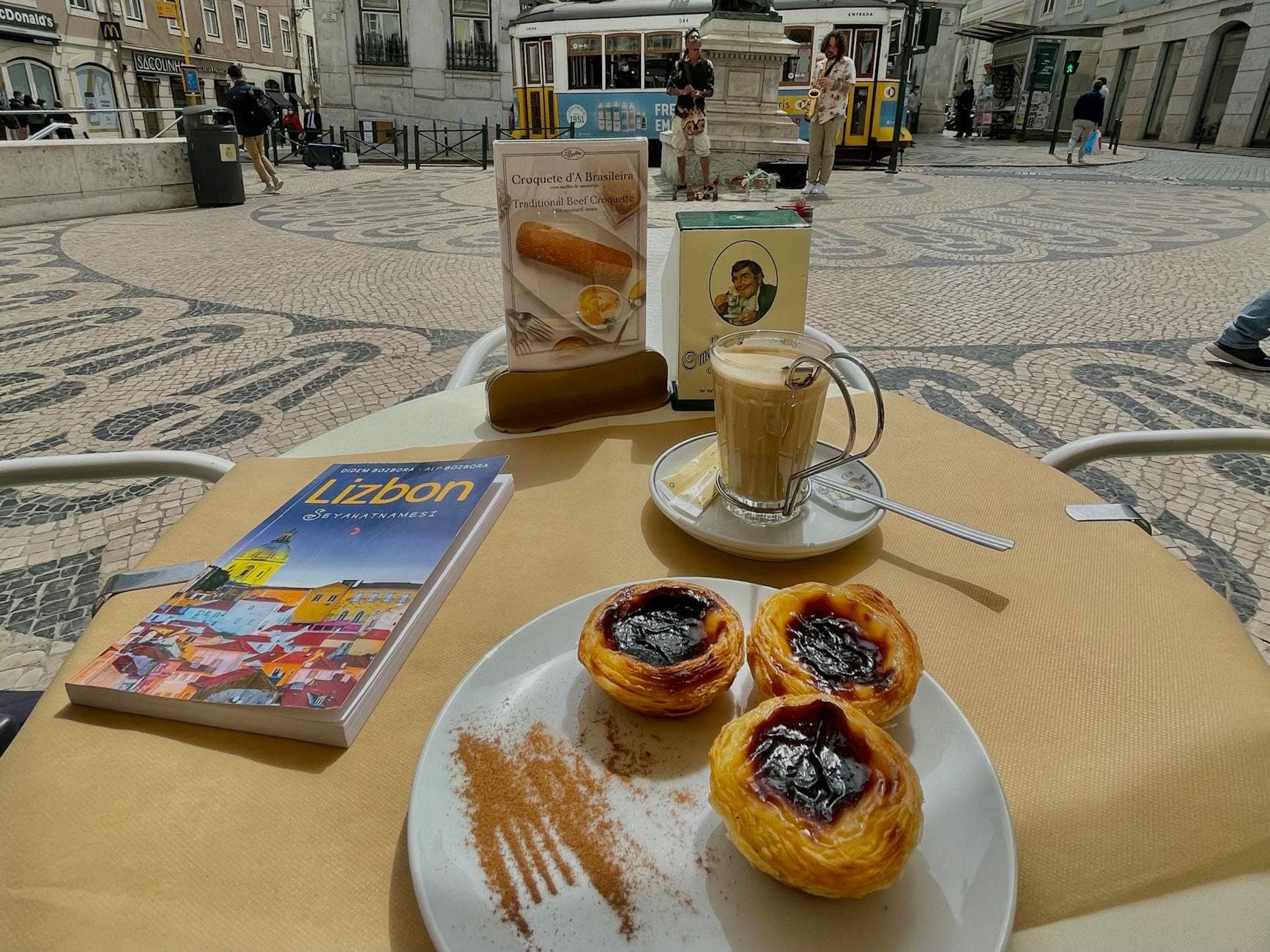 Mesa com três pastéis de nata, um café com leite, um guia de viagem de Lisboa e uma praça da cidade ao fundo com elétrico e edifícios.