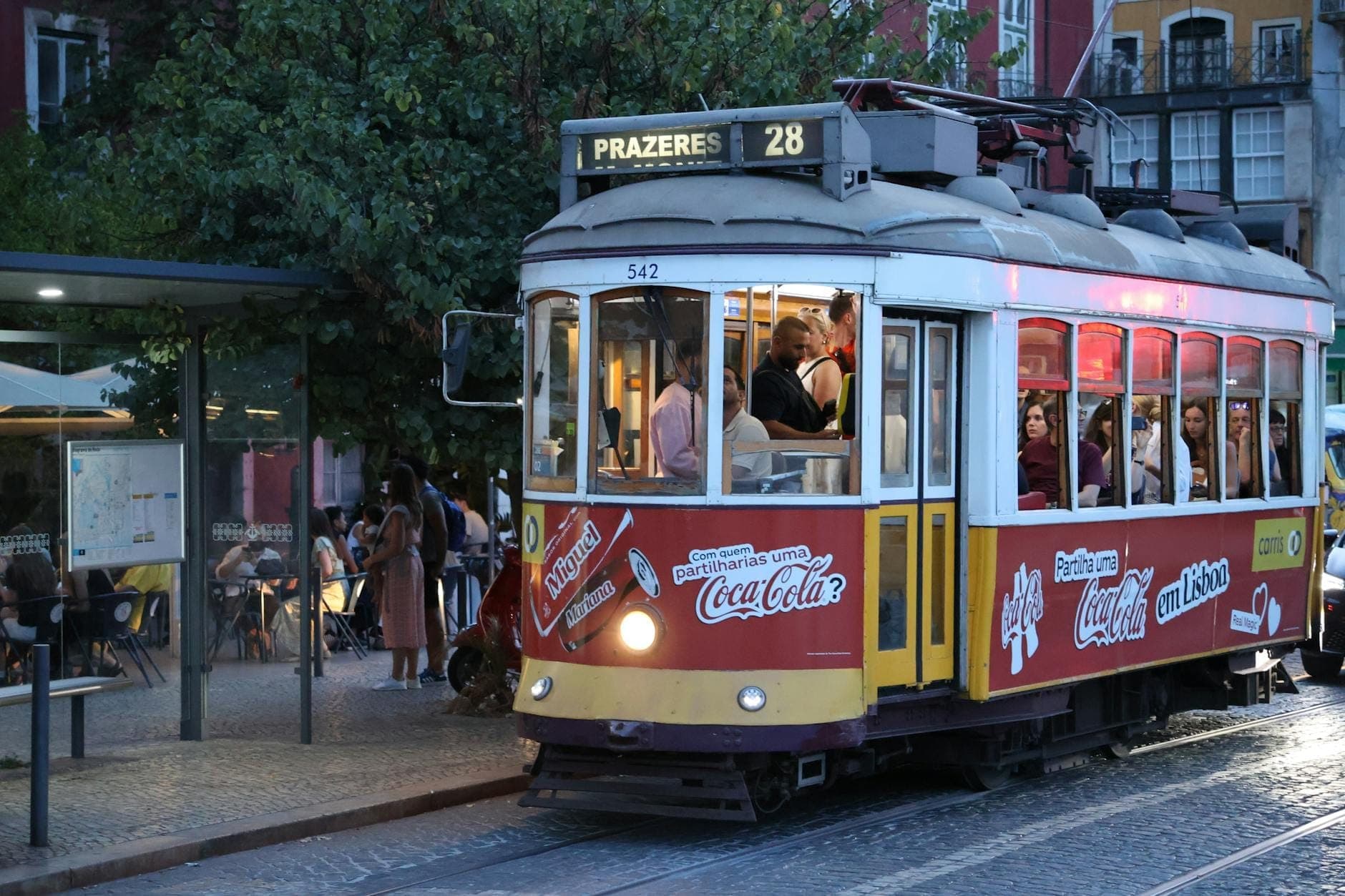 Bonde lotado de Lisboa ao entardecer com muitos passageiros, pessoas esperando na parada e café ao ar livre, ilustrando as multidões e a sazonalidade.