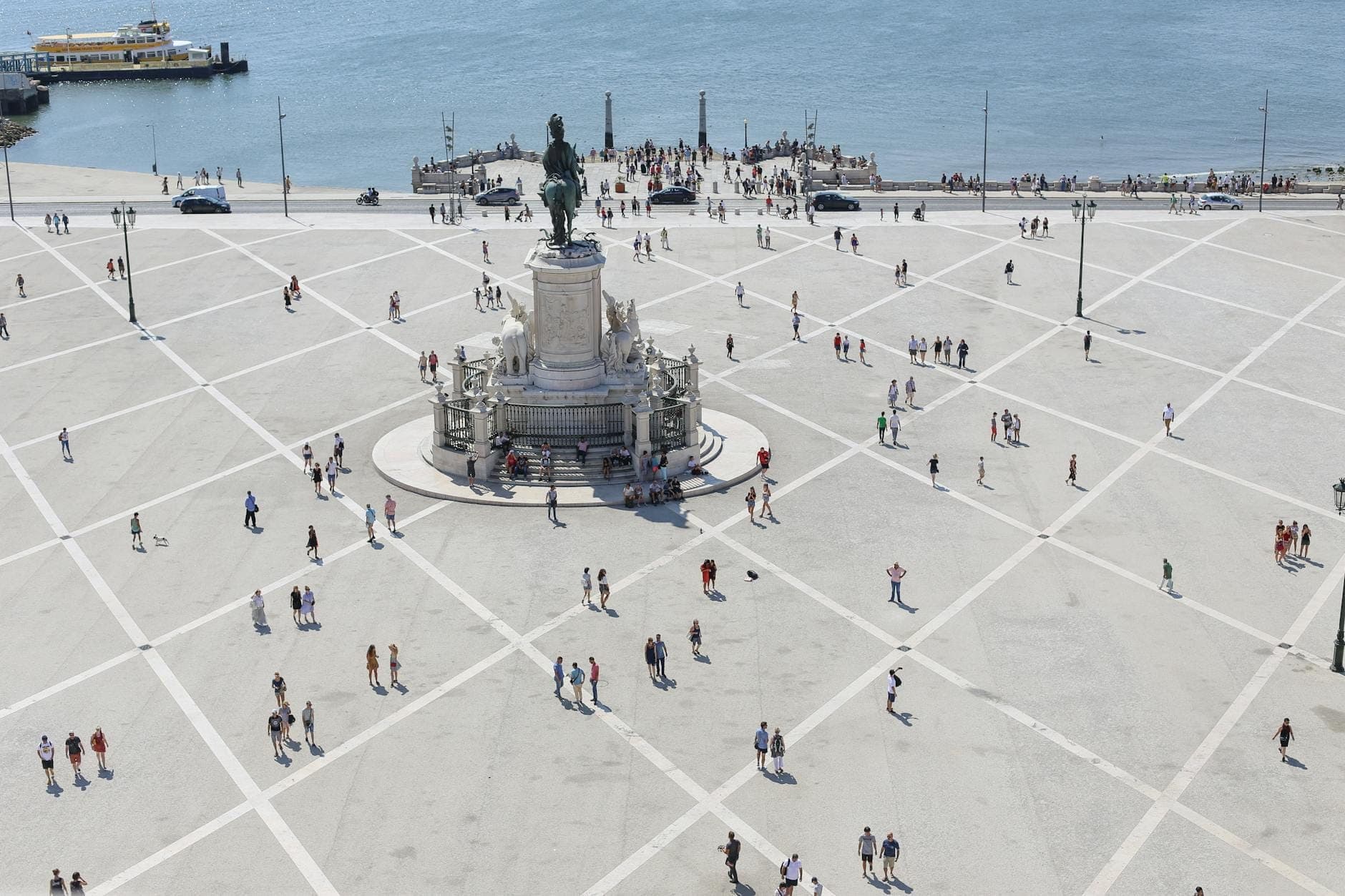 Vista ensolarada da Praça do Comércio de Lisboa, com pessoas aproveitando o espaço ao ar livre à beira do rio Tejo e a estátua central.