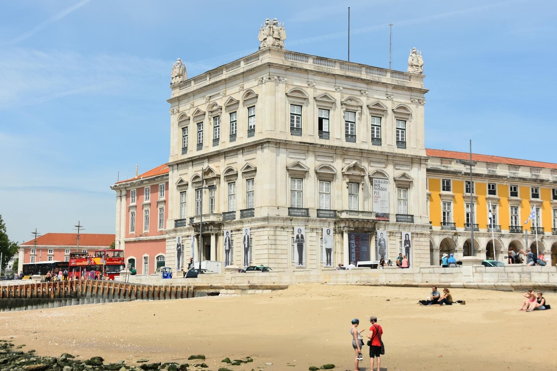 Crianças brincando em uma praia de areia em frente aos coloridos e imponentes prédios de Lisboa em um dia ensolarado, transmitindo um clima perfeito para famílias.