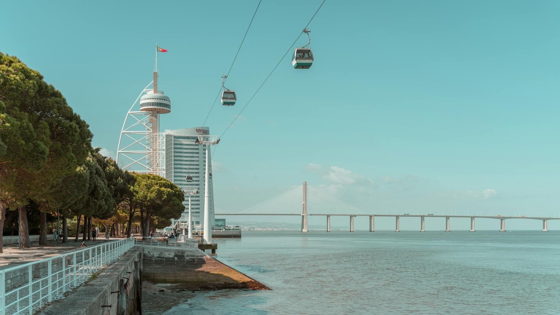 Vista ribeirinha no Parque das Nações com edifícios modernos, teleférico, a torre e a ponte Vasco da Gama, e um passeio ladeado de árvores.