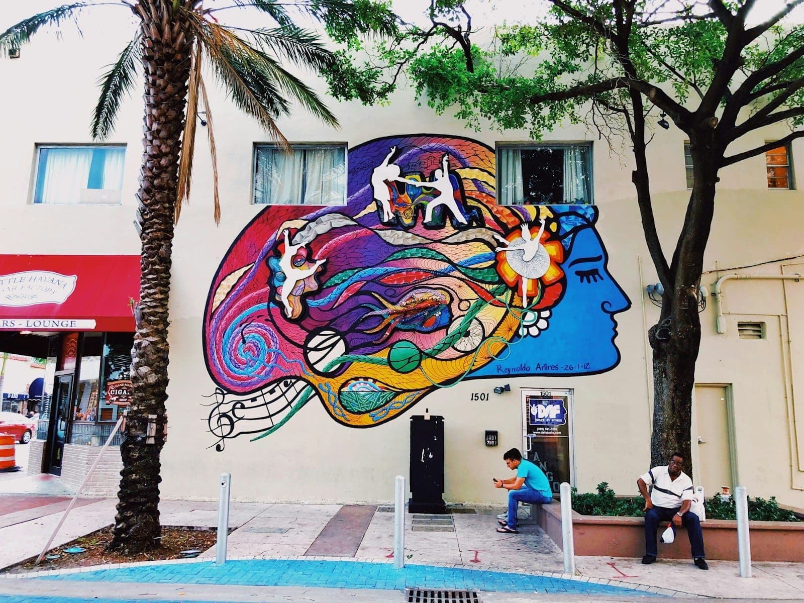 Colorful mural of a woman’s face with vibrant Cuban artistic elements on a street wall, with two people sitting nearby and a palm tree overhead.