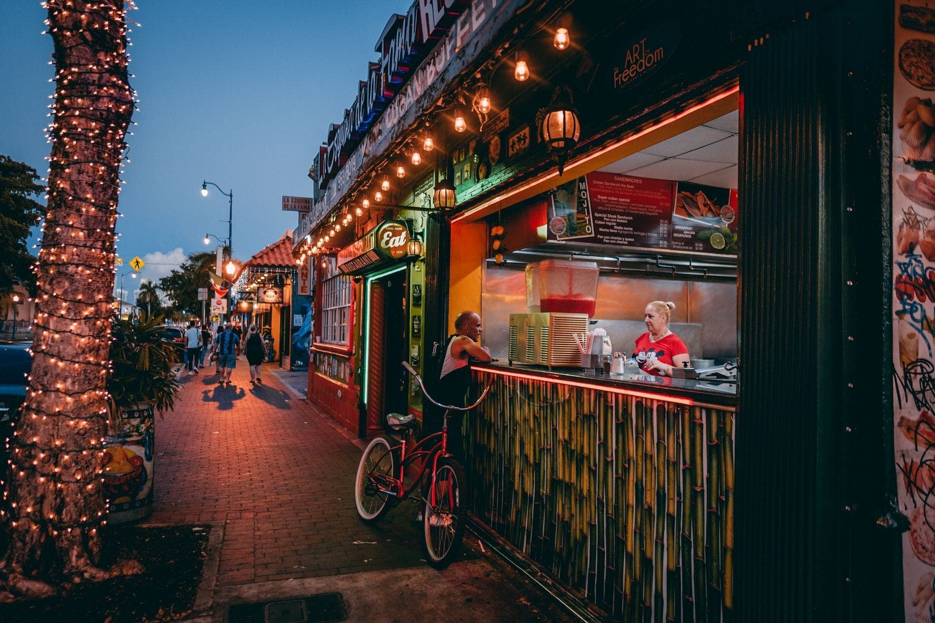 A vibrant Calle Ocho street scene at dusk with people at a walk-up Cuban cafe window, festive string lights, and colorful storefronts in Little Havana, Miami.