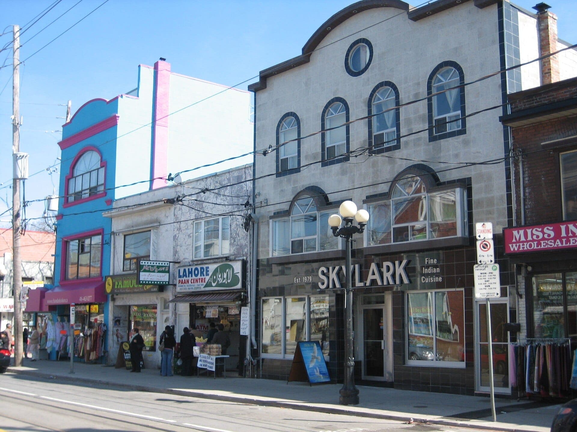 Vista della strada del Gerrard India Bazaar con facciate colorate, negozi di sari, ristoranti e pedoni sotto un cielo azzurro brillante a Toronto.