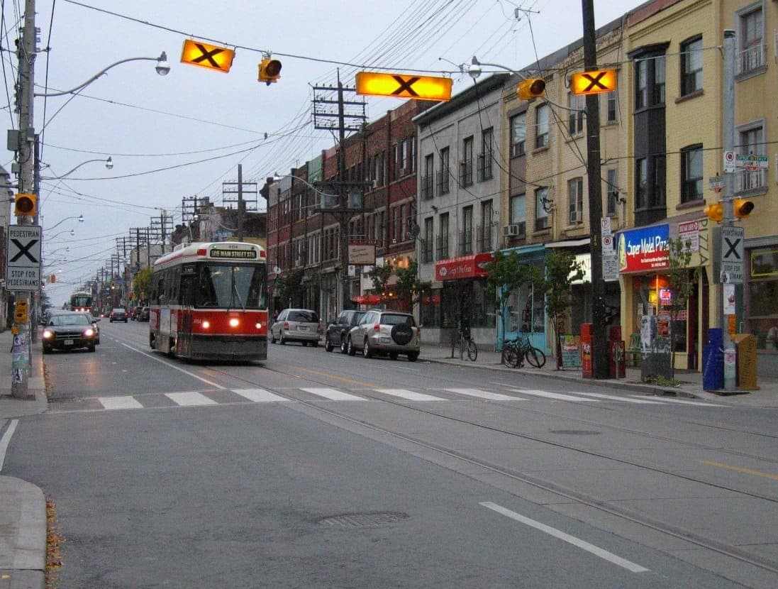 Un tram percorre College Street a Little Italy, Toronto, con negozi colorati e pedoni sotto un cielo coperto e semafori.