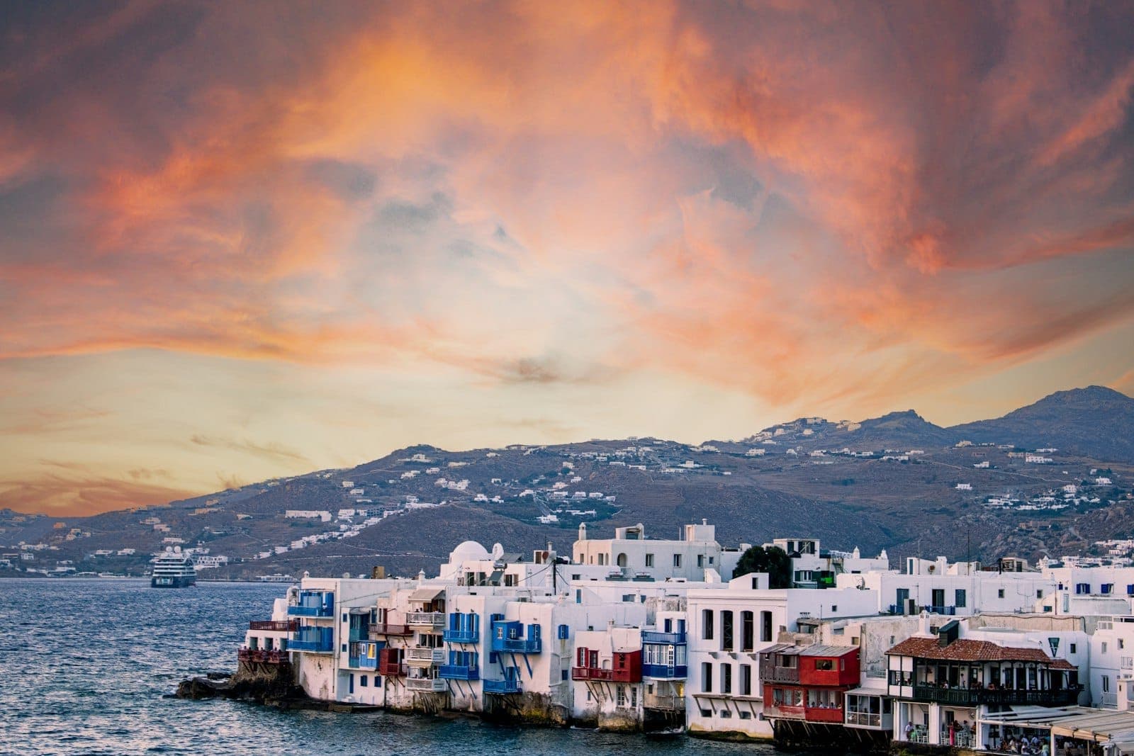 Weiß getünchte Uferhäuser mit bunten Balkonen in Little Venice, Mykonos, vor einem dramatischen Sonnenuntergangshimmel über der Ägäis und fernen Hügeln.