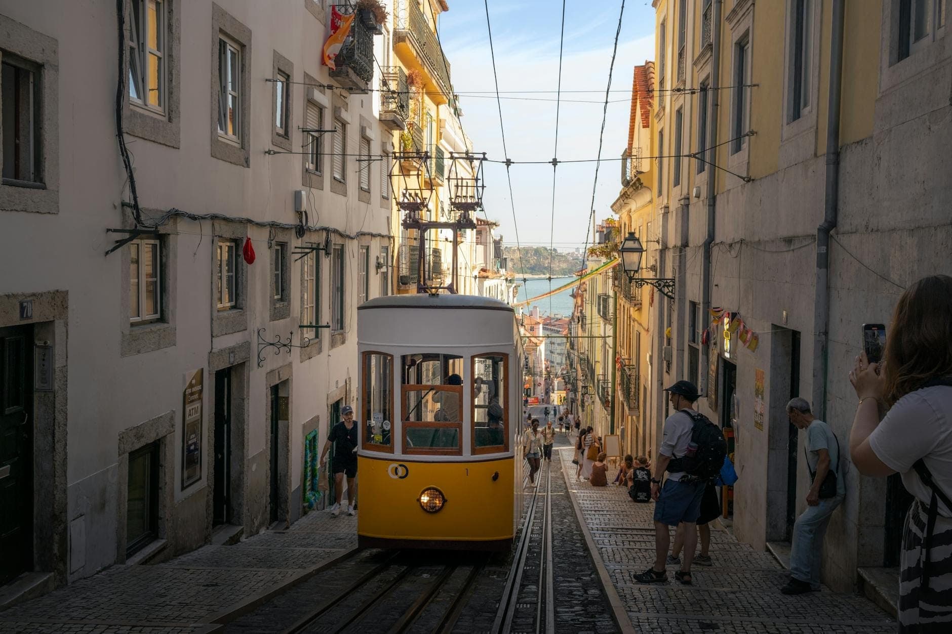 Elétrico amarelo clássico de Lisboa a subir uma íngreme rua de calçada com pessoas a caminhar e o rio visível ao fundo.