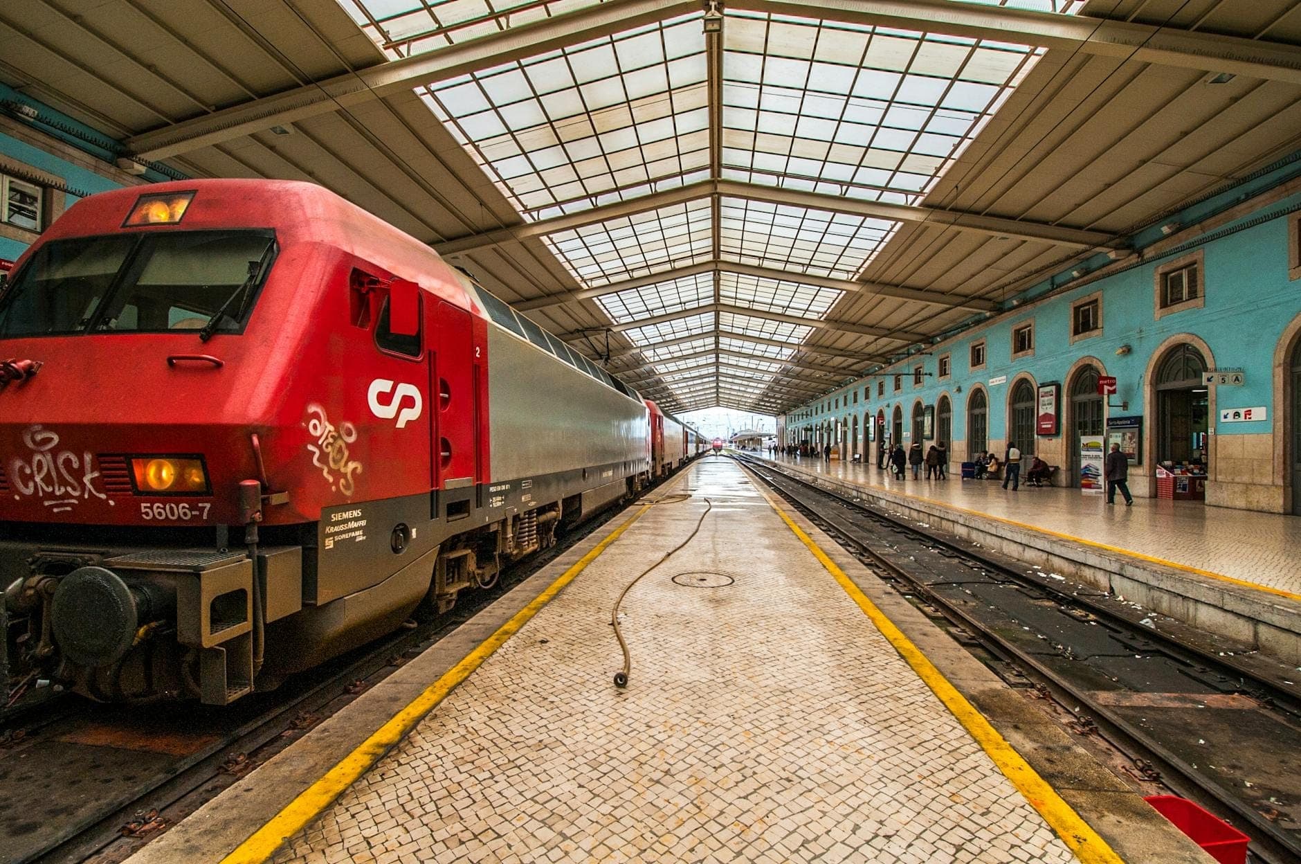 Um trem vermelho em uma estação iluminada com teto de vidro, paredes azuis e pessoas caminhando pela plataforma, evocando o início de uma jornada de viagem.