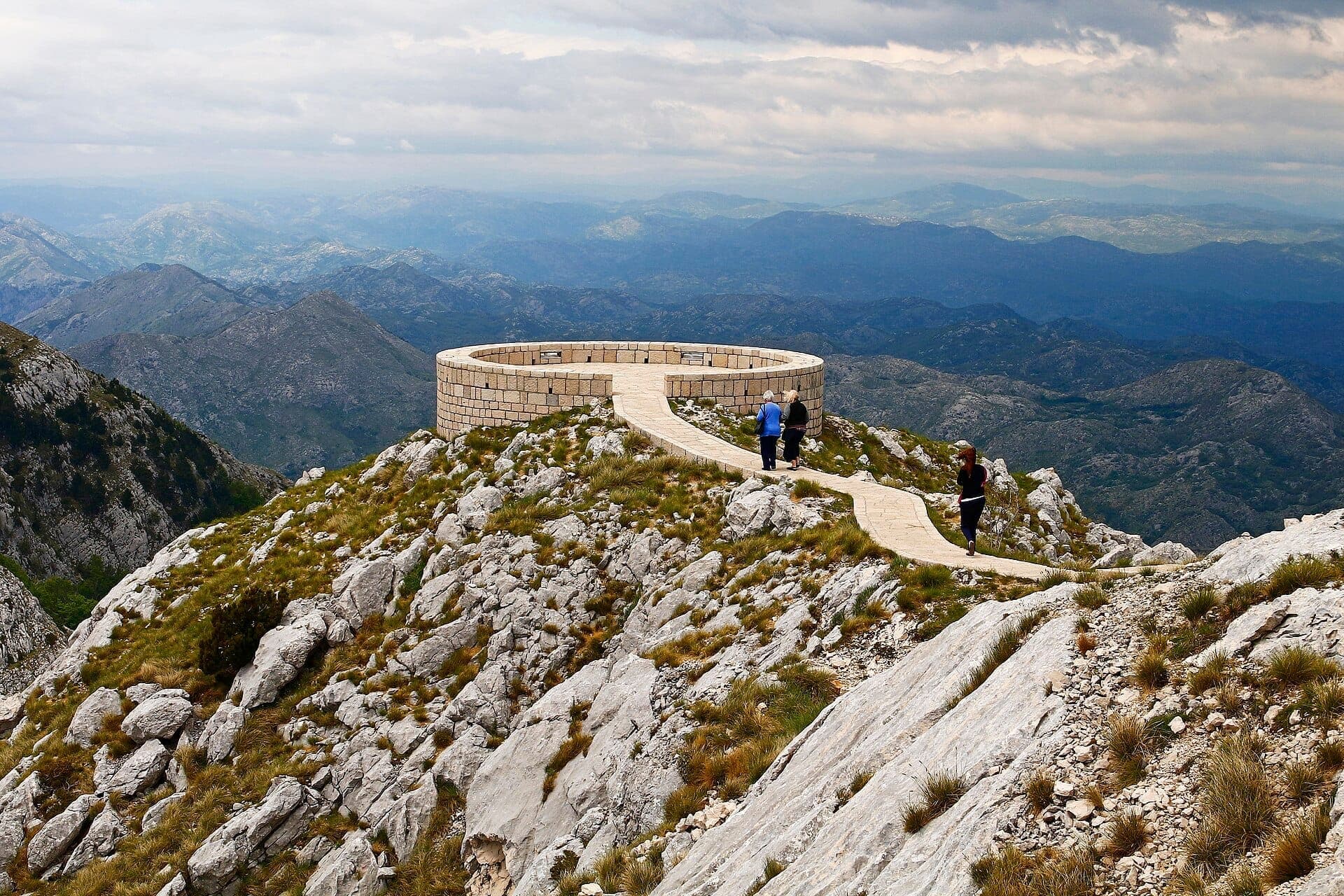 Belvédère circulaire en pierre au sommet du mont Lovćen, entouré de pics rocheux spectaculaires et d'un ciel nuageux, avec trois visiteurs s'approchant par un sentier étroit.