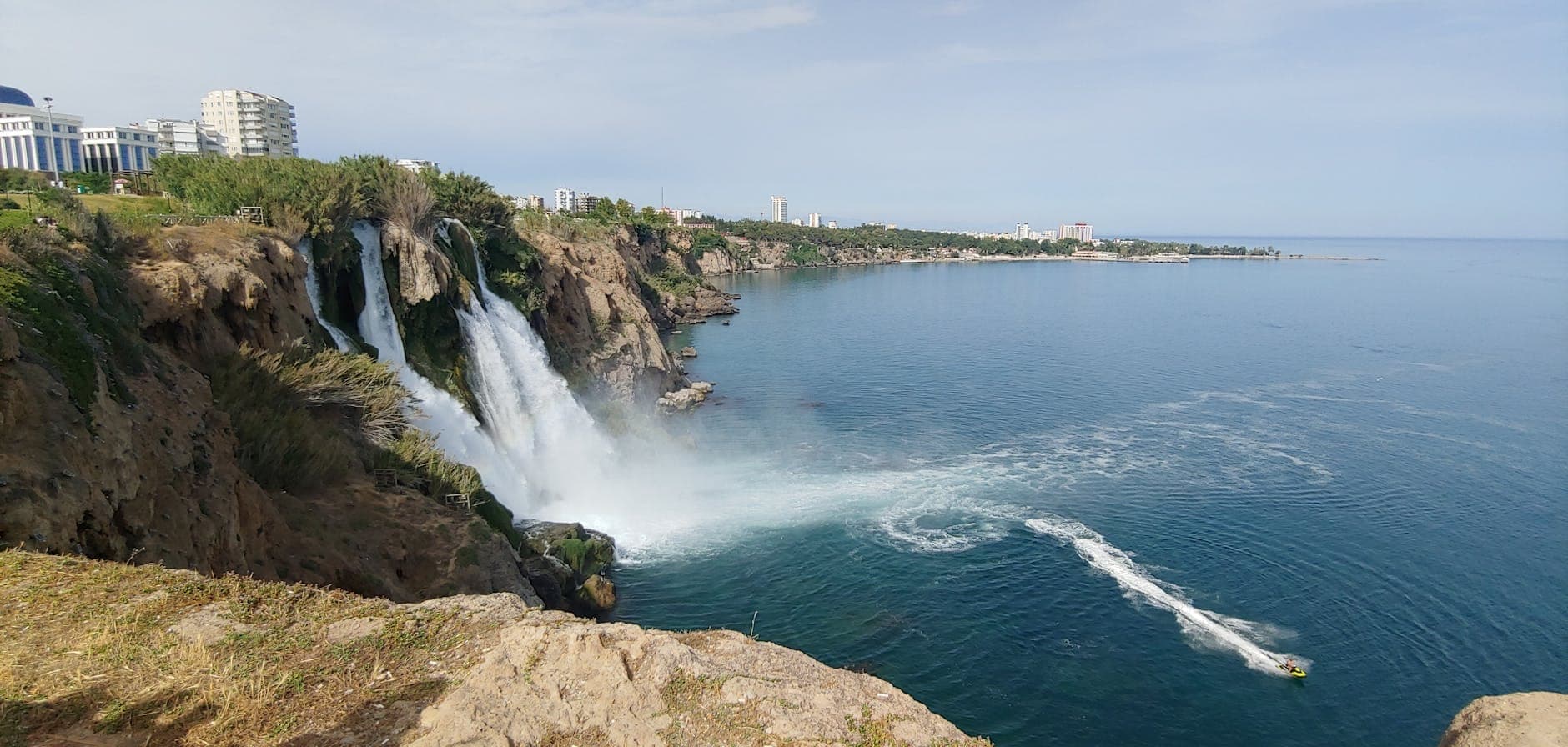 Weitwinkelpanorama des unteren Düden-Wasserfalls, der von einer Klippe ins Mittelmeer stürzt – mit umliegenden Gebäuden, klarem Himmel und einem Boot auf dem Wasser darunter.