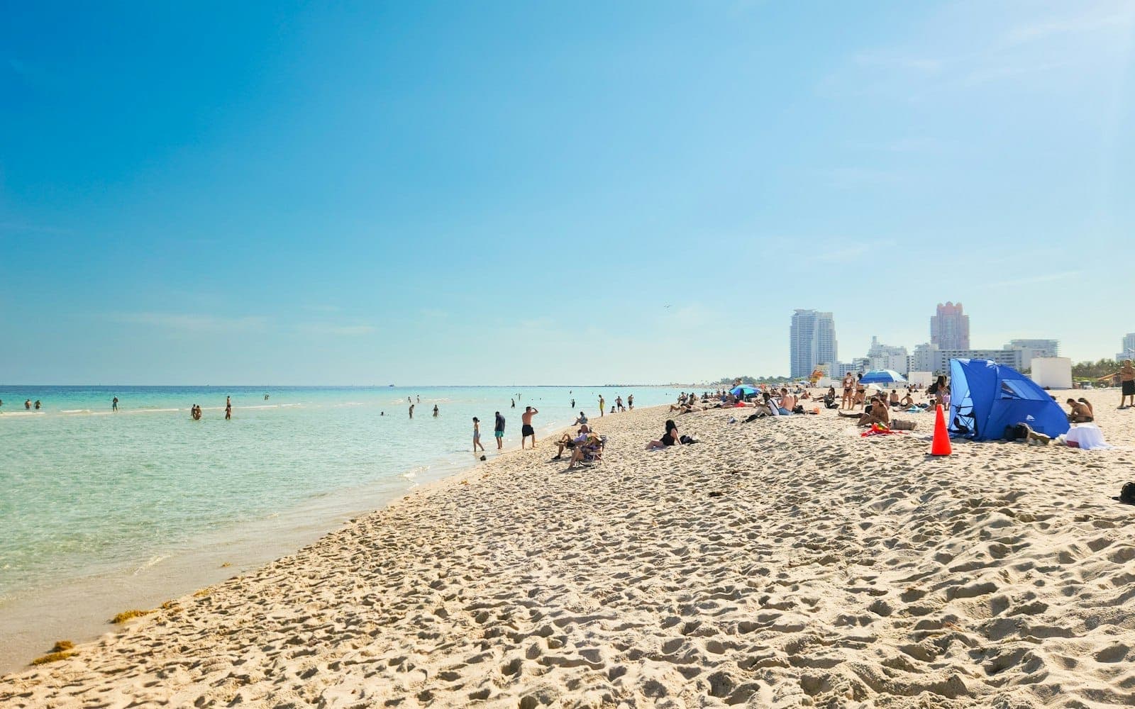 A wide view of Lummus Park Beach showing clear blue sky, turquoise water, soft sandy shore, and people relaxing and swimming with Miami Beach skyline in the distance.
