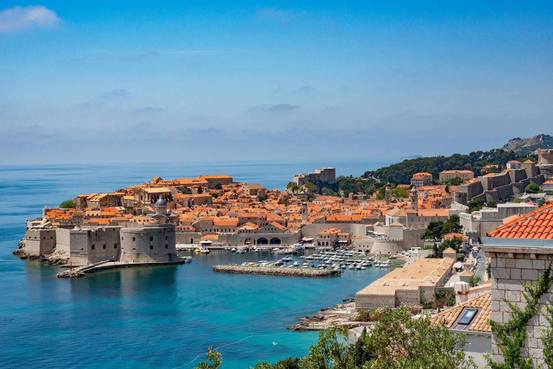 Panoramablick auf die Steinmauern und den Hafen der Altstadt von Dubrovnik, mit klarem blauem Meer und historischen Gebäuden unter einem strahlenden Himmel.
