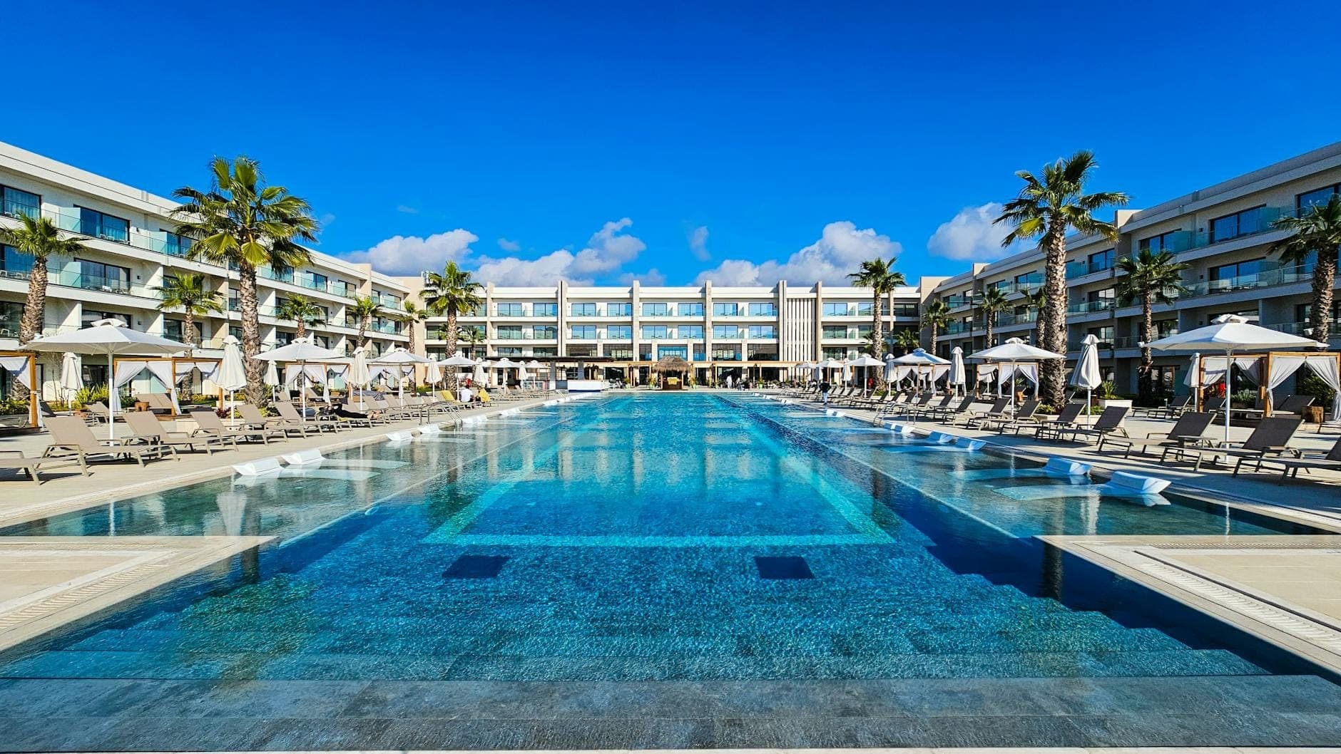 Hôtel moderne et luxueux en bord de mer avec grande piscine, chaises longues, palmiers et ciel bleu, représentant un hébergement haut de gamme à Rhodes.