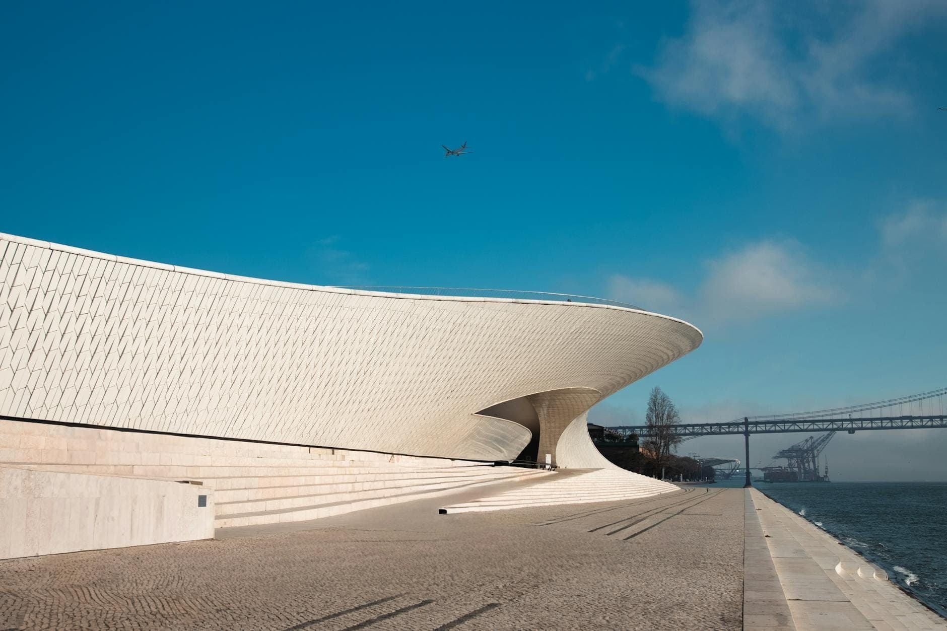A icônica galeria de forma ondulada do MAAT, revestida de azulejos brancos, à beira do Tejo sob um céu azul e sol forte, com a ponte ao fundo.